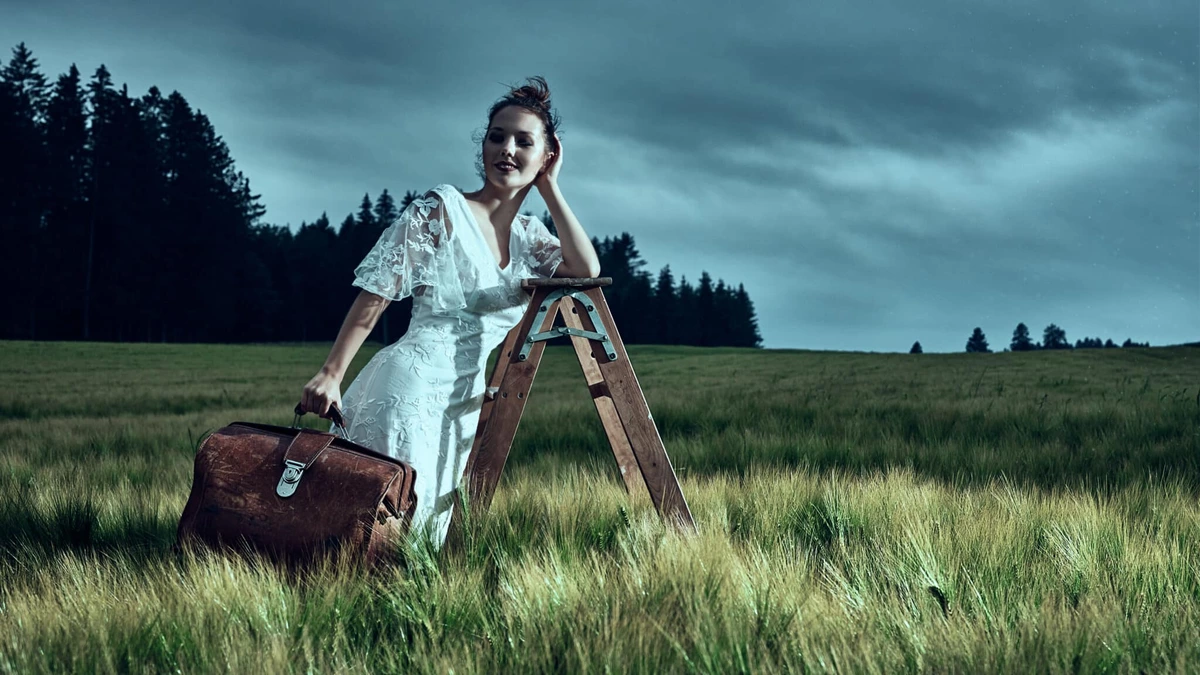 Eliane en robe blanche appuyée sur un escabeau dans un champ, valise en cuir, ciel d'orage