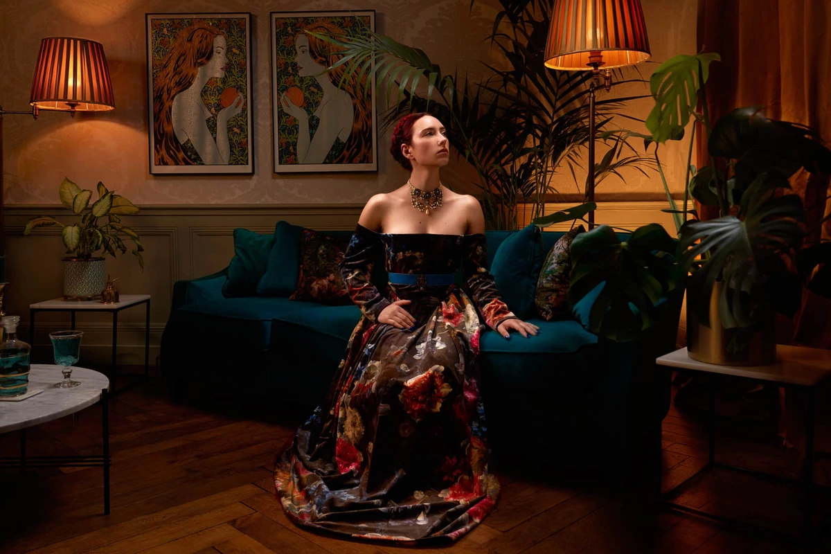 Maeva en robe fleurie, assise dans un salon rétro aux lampes chaudes, plantes et tableaux