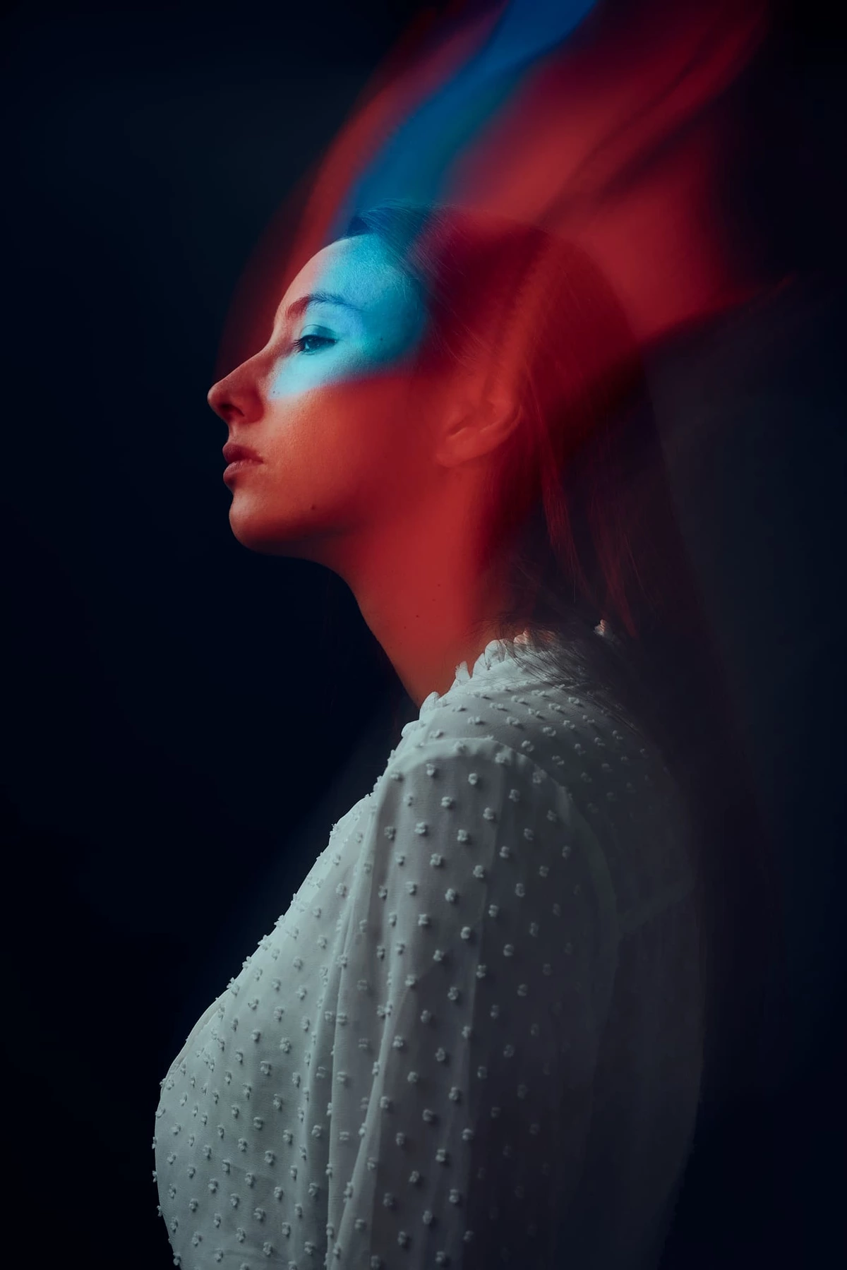 Maeva de profil, visage éclairé de bleu et cheveux traînés de rouge, sur fond noir