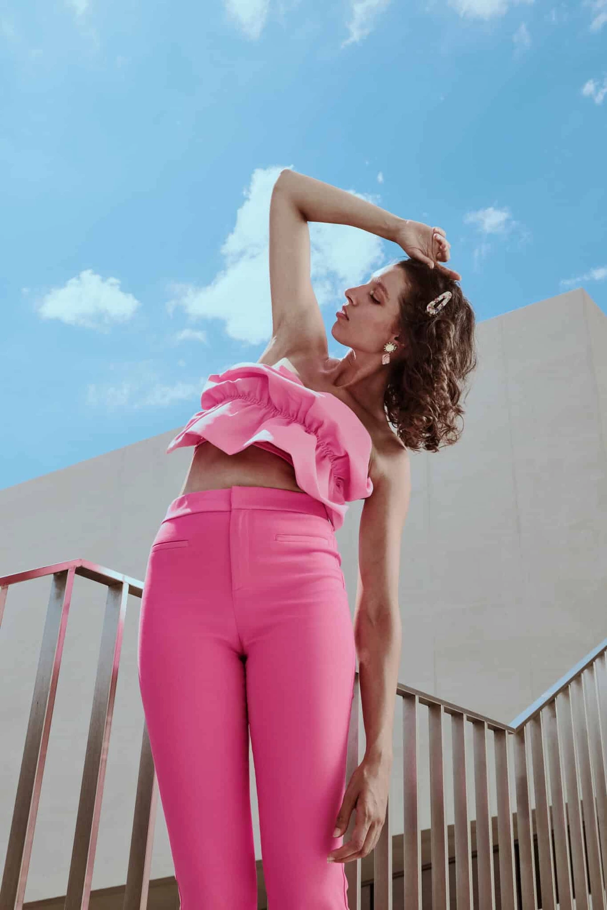 Camille en ensemble rose, bras levé, sur un escalier en béton sous un ciel bleu nuageux