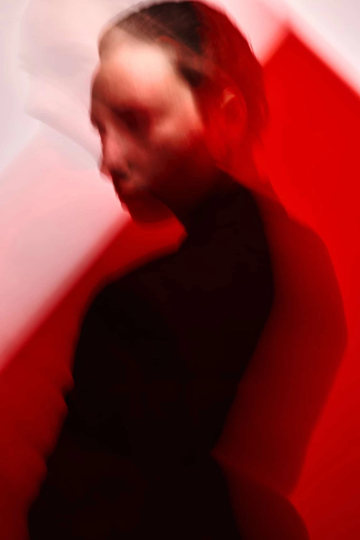 Maeva floutée par le mouvement, tête baissée, en veste noire devant un aplat rouge vif