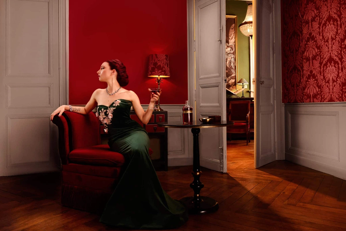 Maeva en robe verte, assise sur un fauteuil rouge dans un salon aux murs rouges, un verre à la main