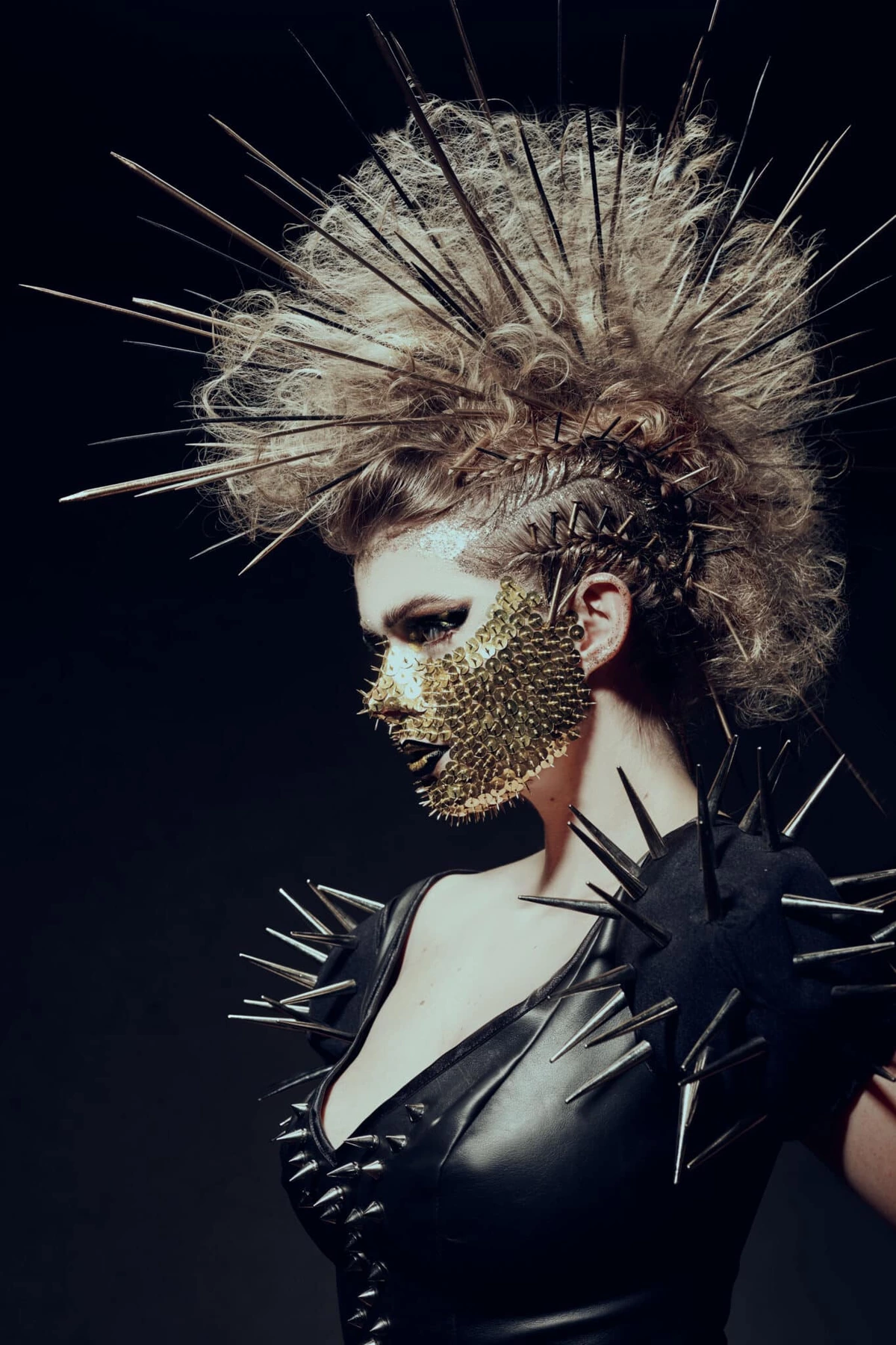 Audrey de profil, coiffe de pointes et masque doré, tenue en cuir à pointes, fond noir