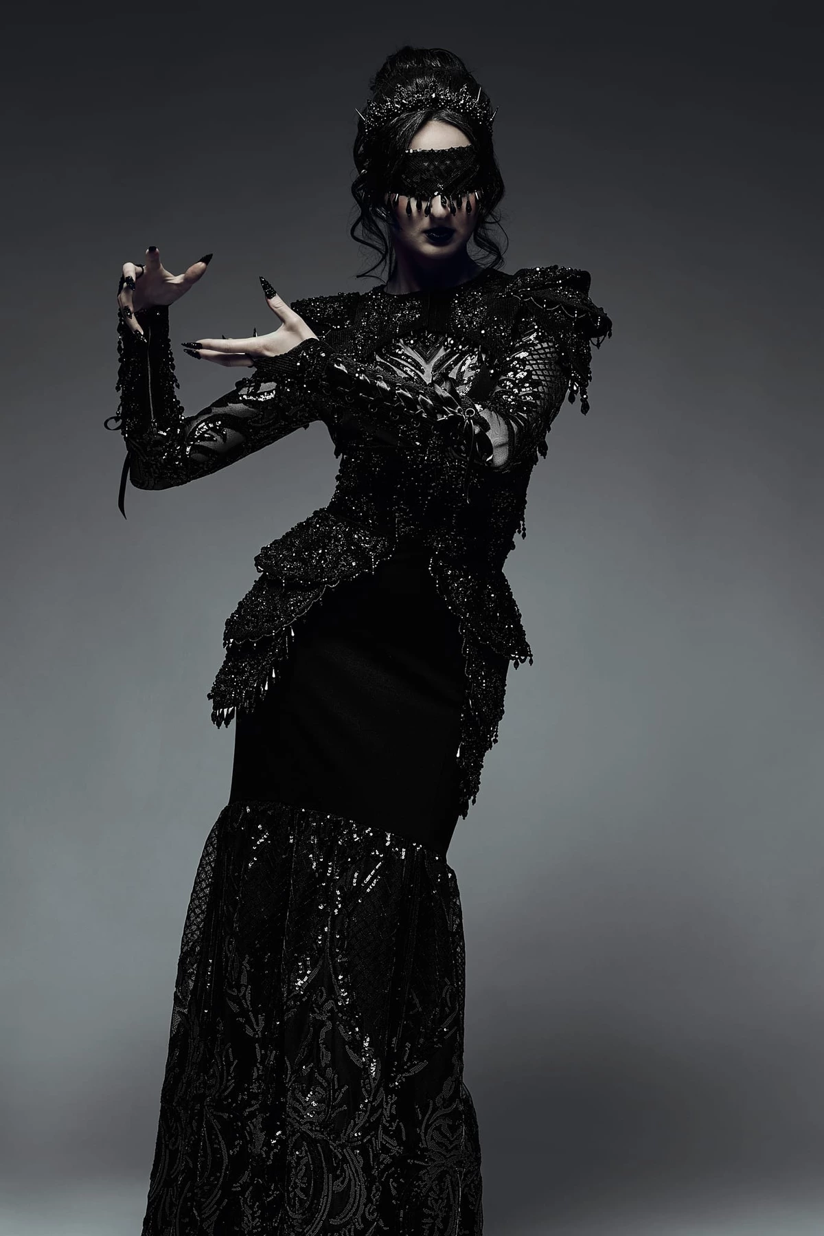Lucie en robe noire ornée et masque, posture théâtrale, mains levées sur fond gris