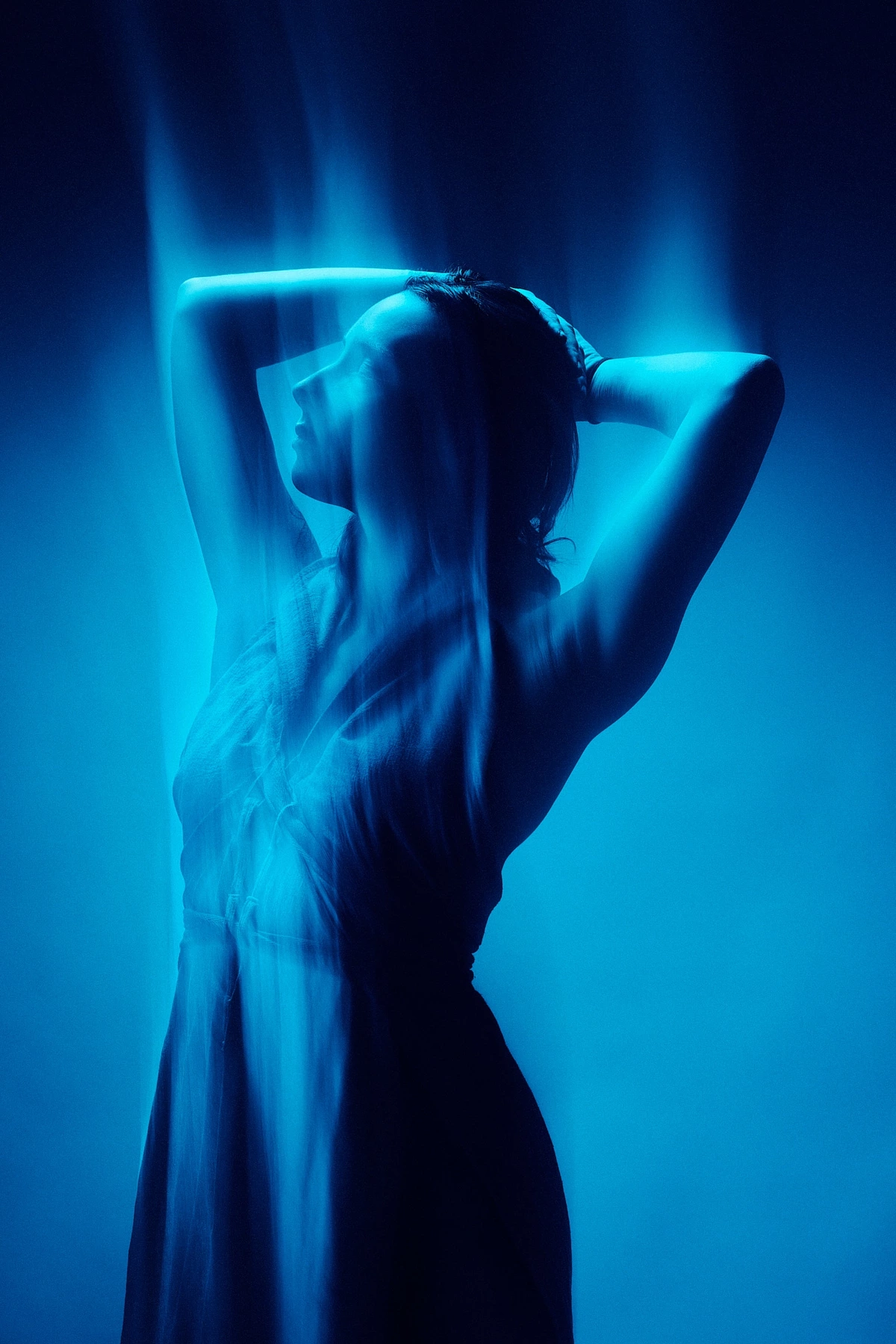 Maeva in a flowing dress, arms raised above her head, light trails in a blue monochrome