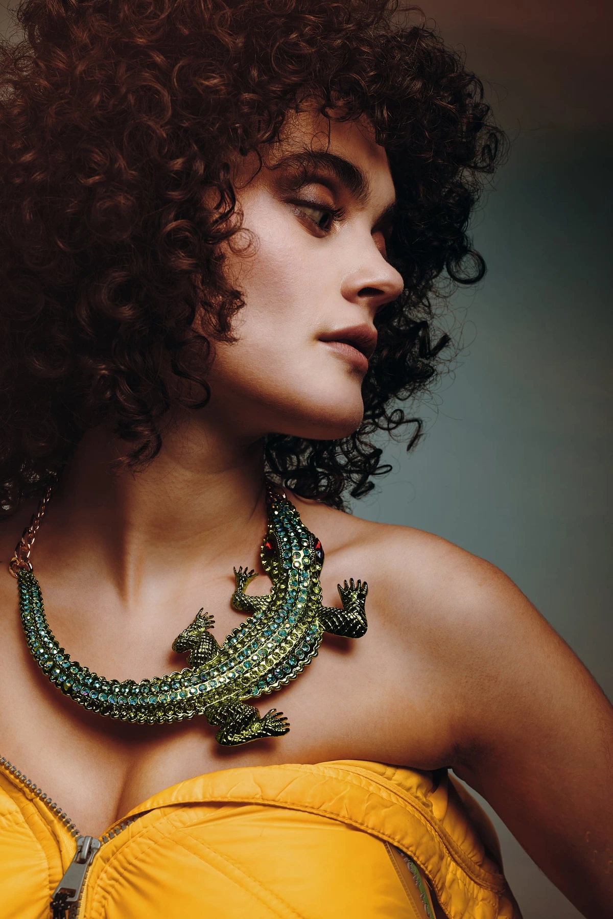 Céline de profil, cheveux bouclés, collier lézard vert serti et veste jaune, fond gris