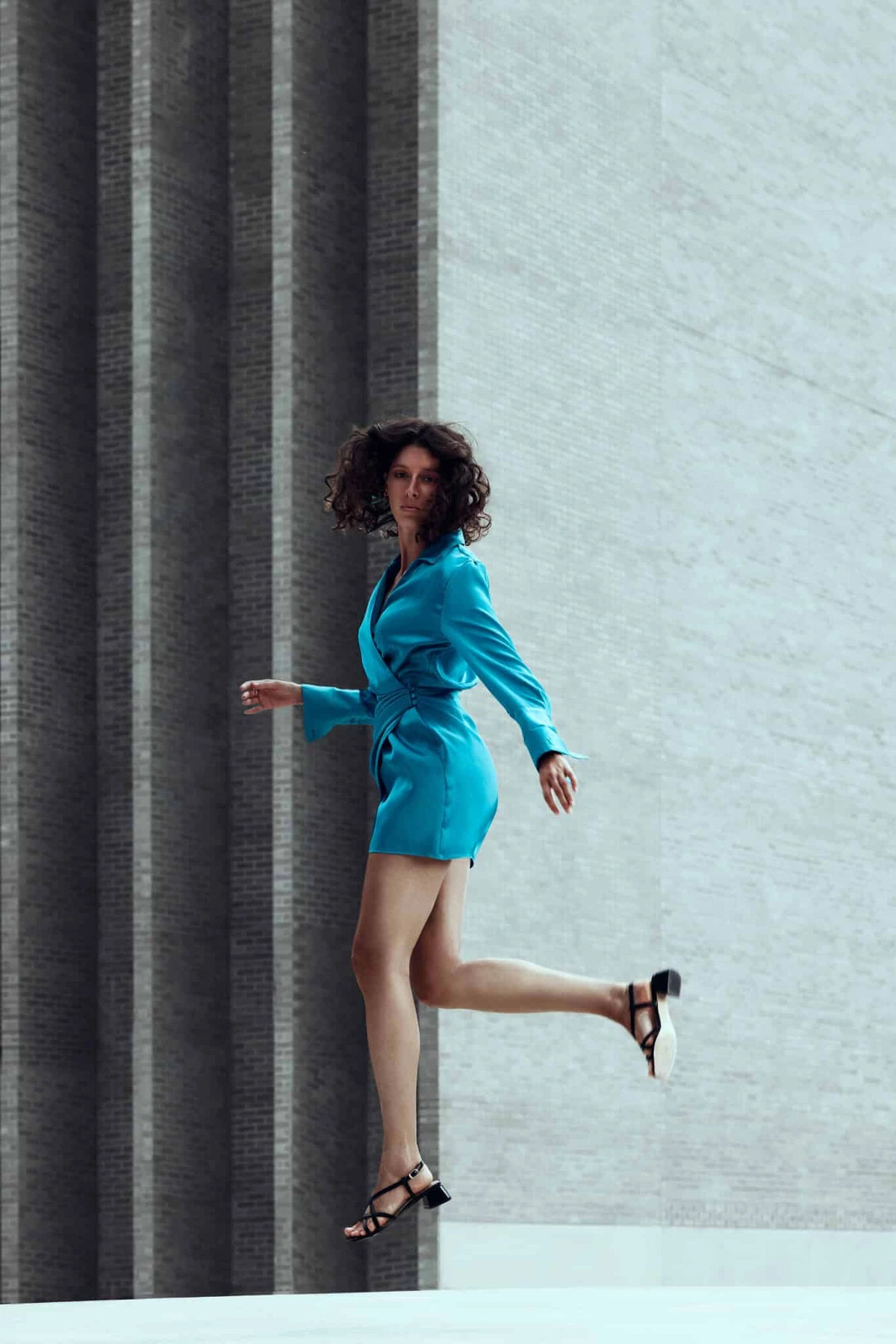 Camille en robe bleue sautant sur une jambe devant un mur clair et des colonnes grises