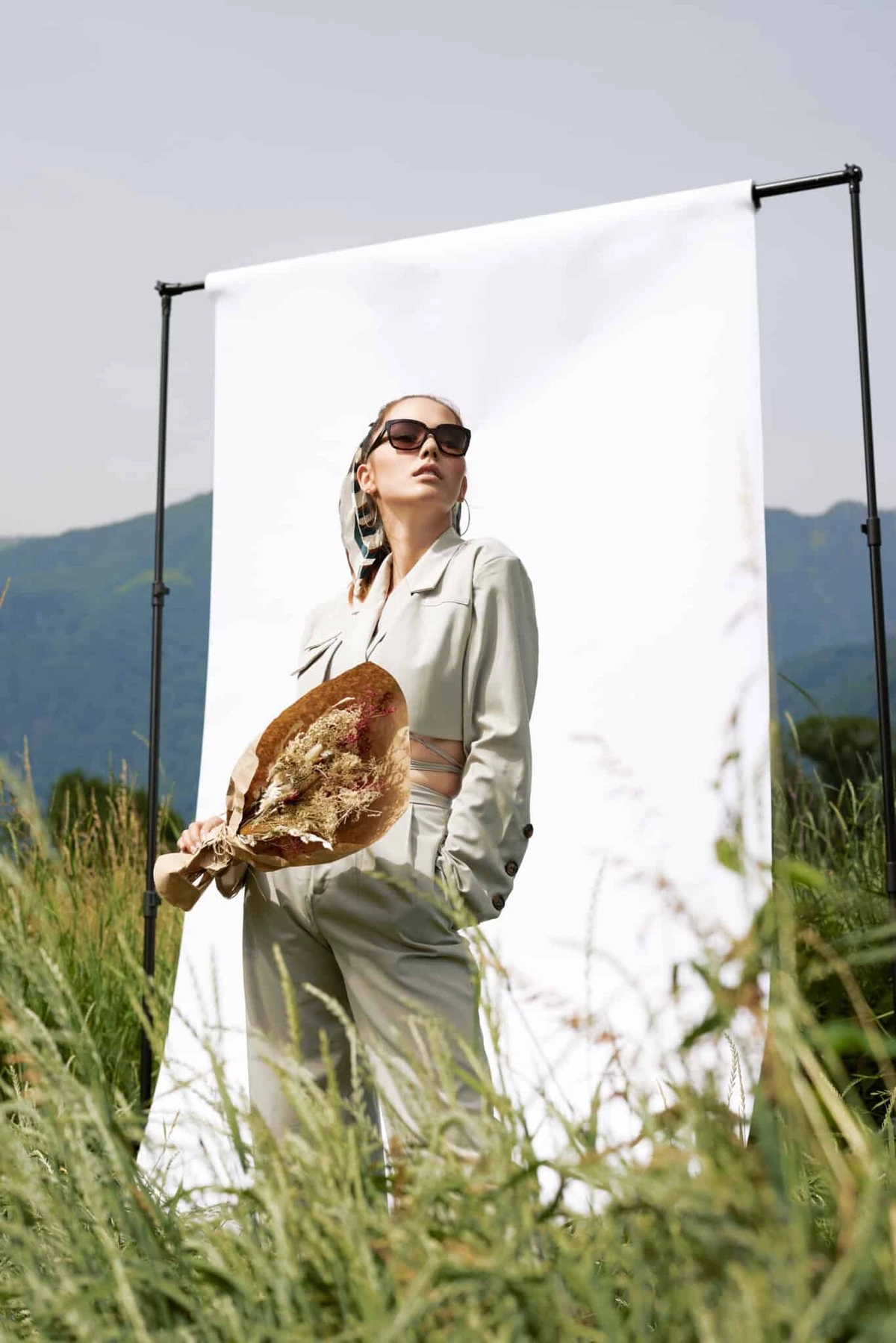 Eliane en tailleur clair et lunettes de soleil devant un fond blanc tendu dans les hautes herbes