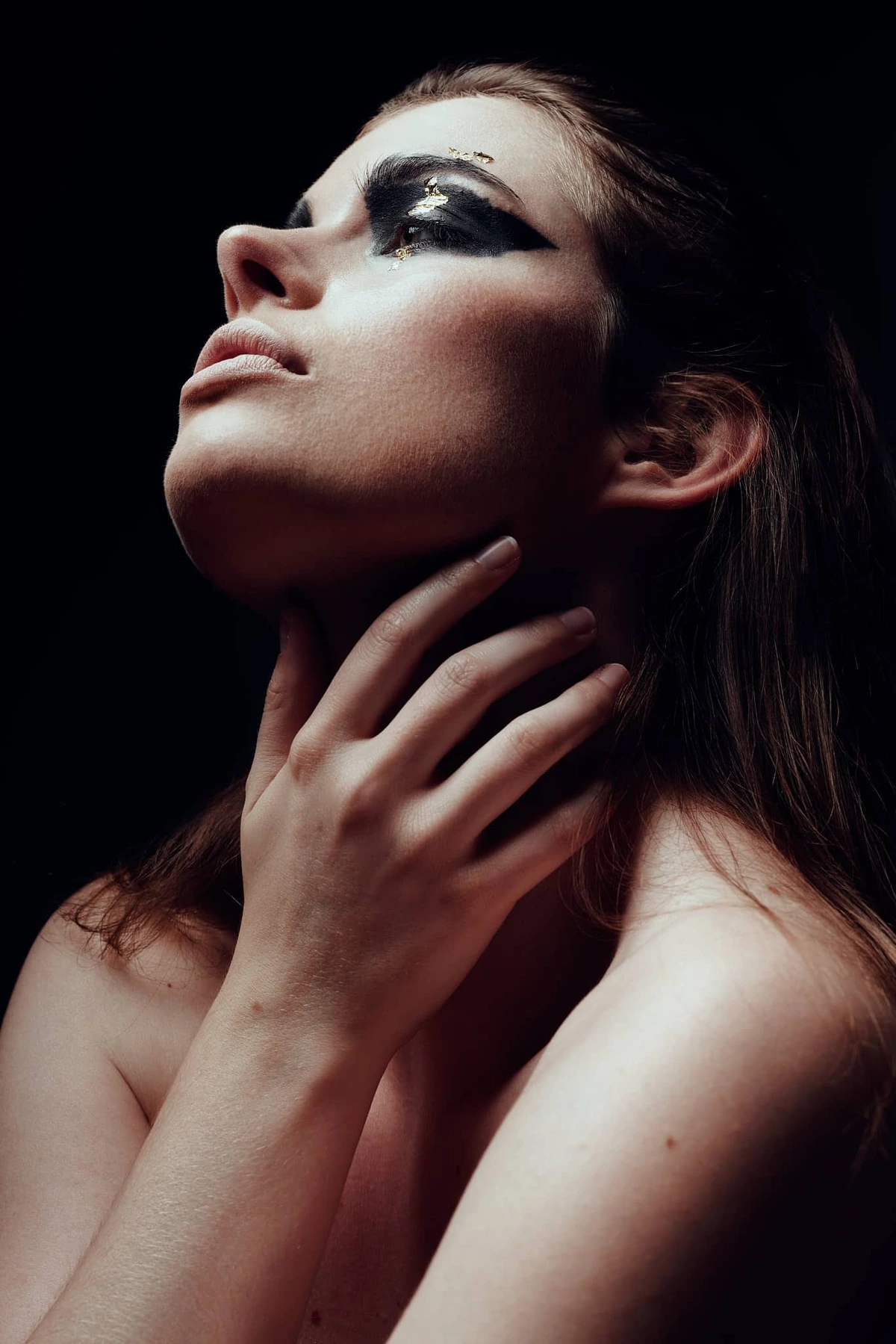 Audrey tête levée, maquillage noir intense, main sur le cou, épaules nues, fond noir