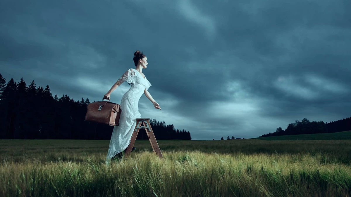 Eliane en robe blanche monte sur un escabeau dans un champ, valise à la main, ciel bleu orageux