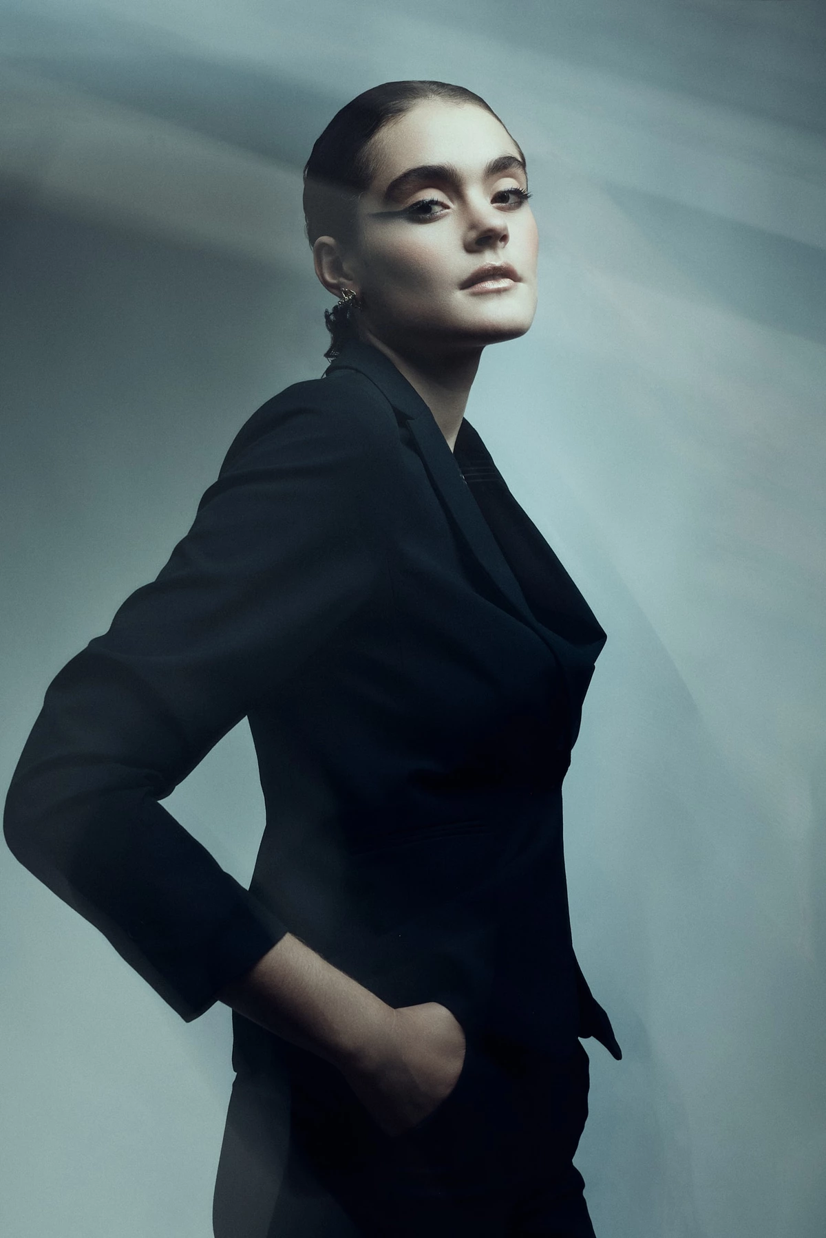 Céline L., cheveux tirés et main dans la poche, en tailleur noir, regard de côté sur fond bleu-gris