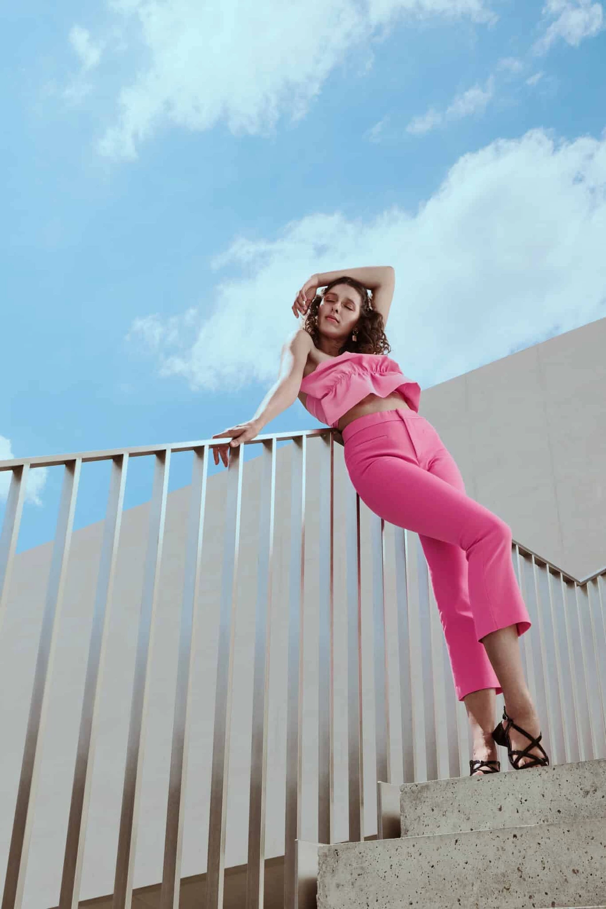 Camille en ensemble rose appuyée à une rambarde d'escalier, vue en contre-plongée, ciel bleu