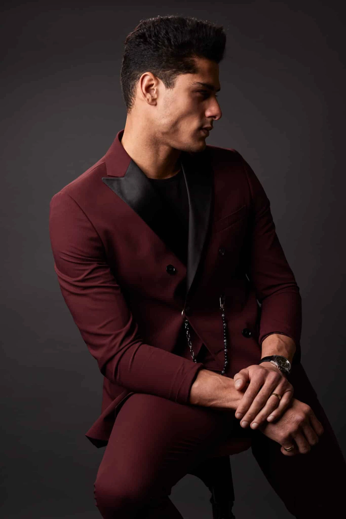 Hiago en costume bordeaux, assis de profil, mains jointes sur le genou, fond sombre