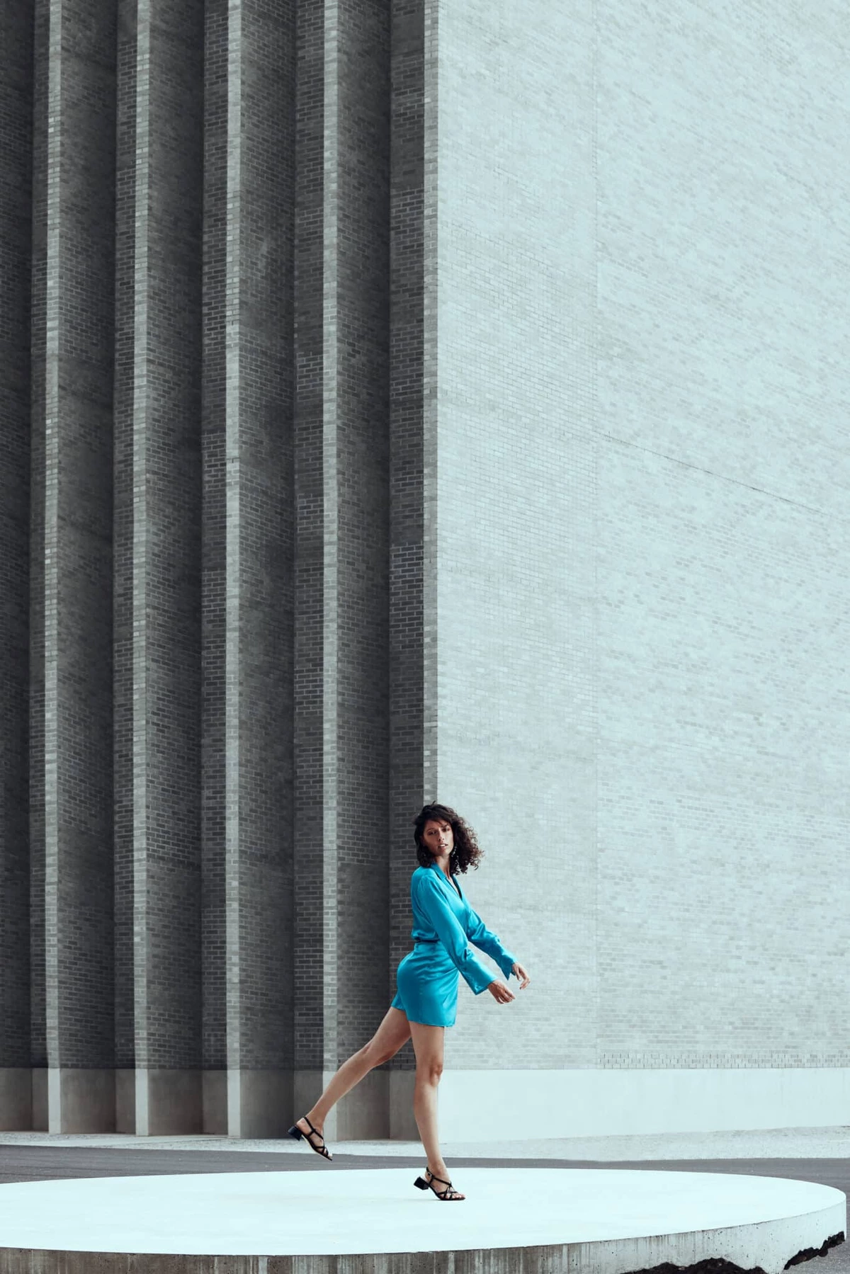 Camille en robe bleue marchant sur une estrade ronde devant un grand mur à colonnes