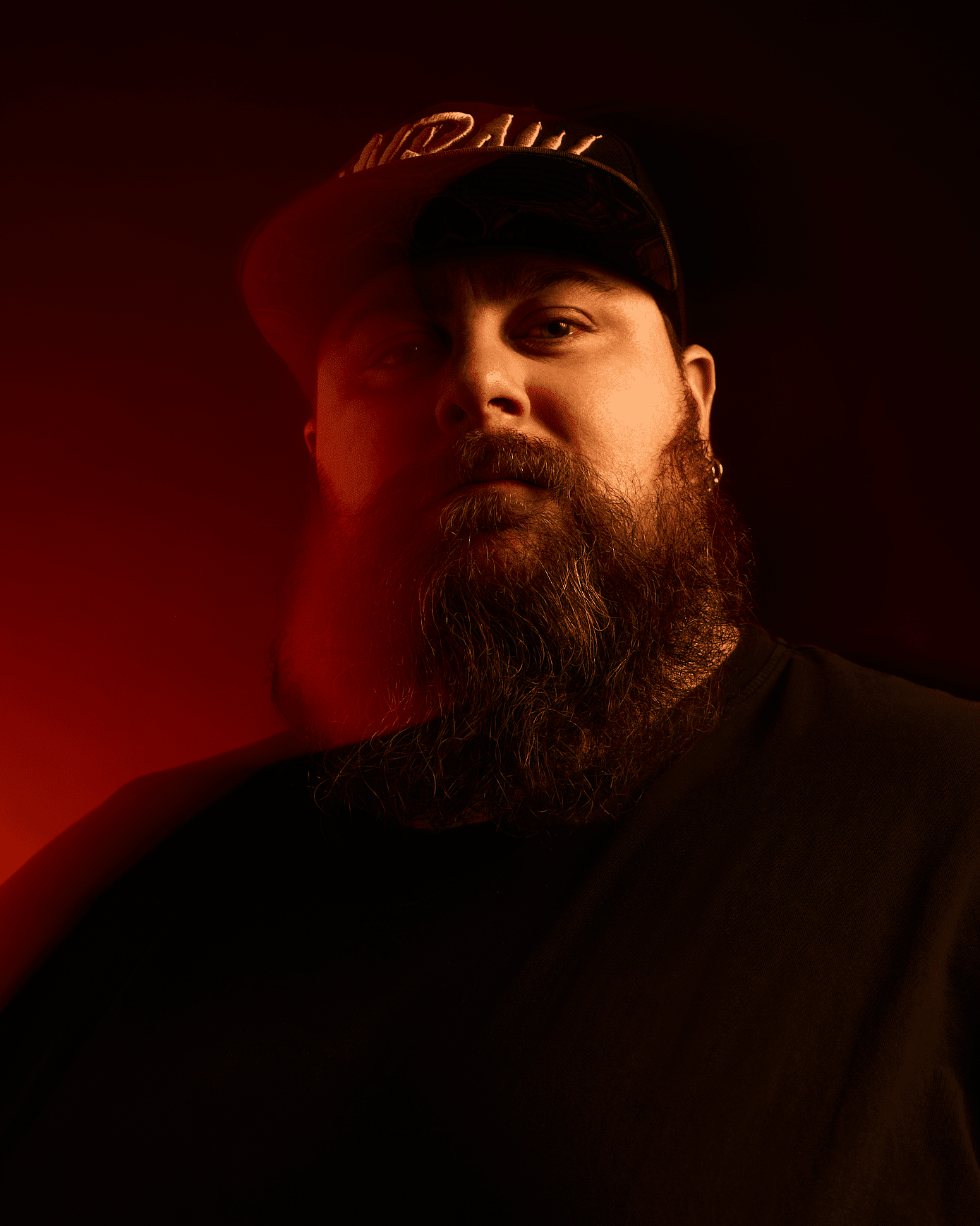 Un homme barbu, vêtu d'une chemise et d'un chapeau noirs, est le point de mire principal sur un fond sombre, un éclairage rouge dramatique mettant en valeur son visage et sa barbe.