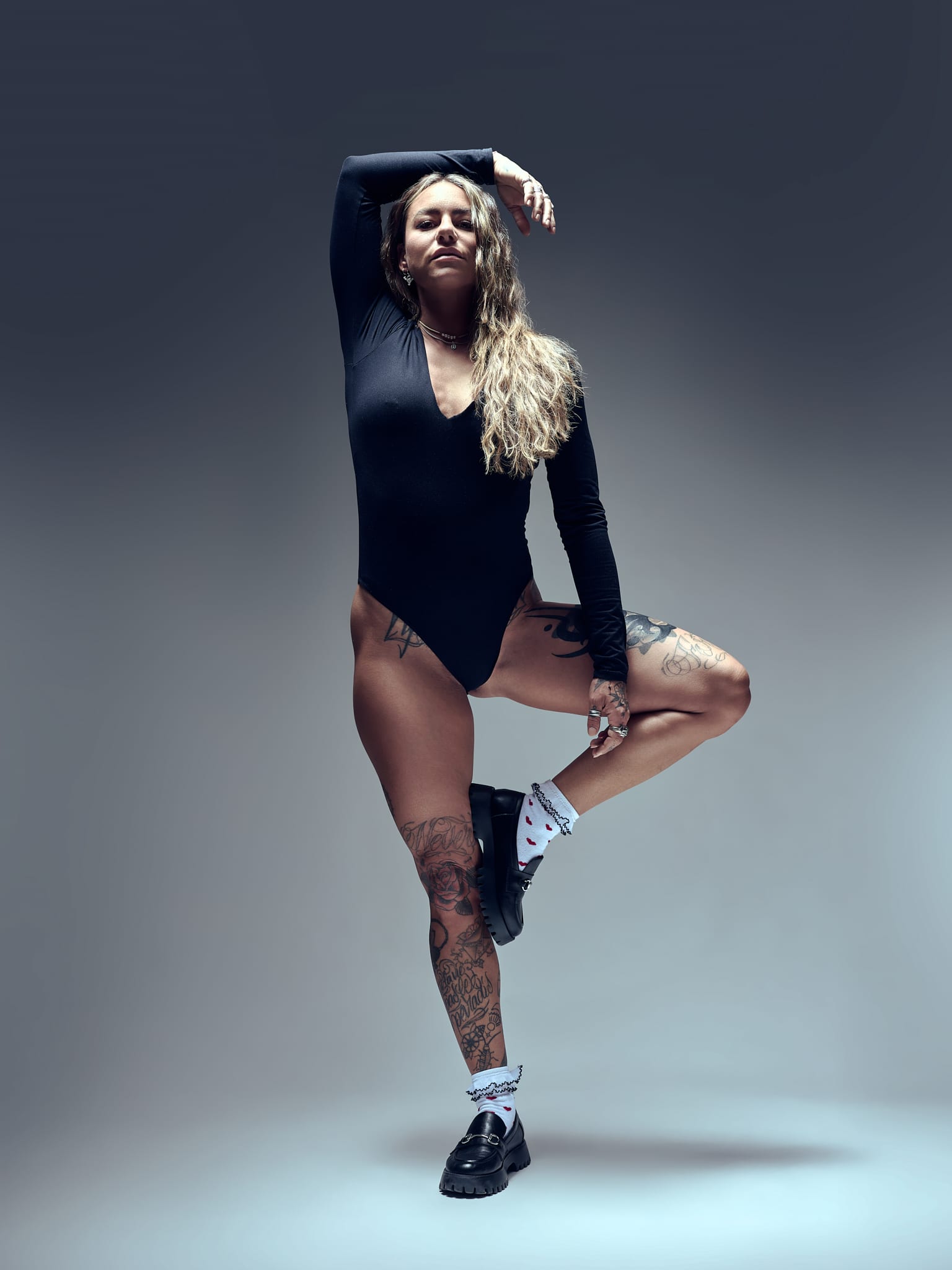 Une femme aux cheveux longs et ondulés pose avec assurance dans un body noir, des chaussettes blanches et des chaussures noires. Elle se tient debout sur une jambe, l'autre pliée, le bras levé au-dessus de la tête, montrant les tatouages sur ses bras et ses jambes sur un fond neutre.