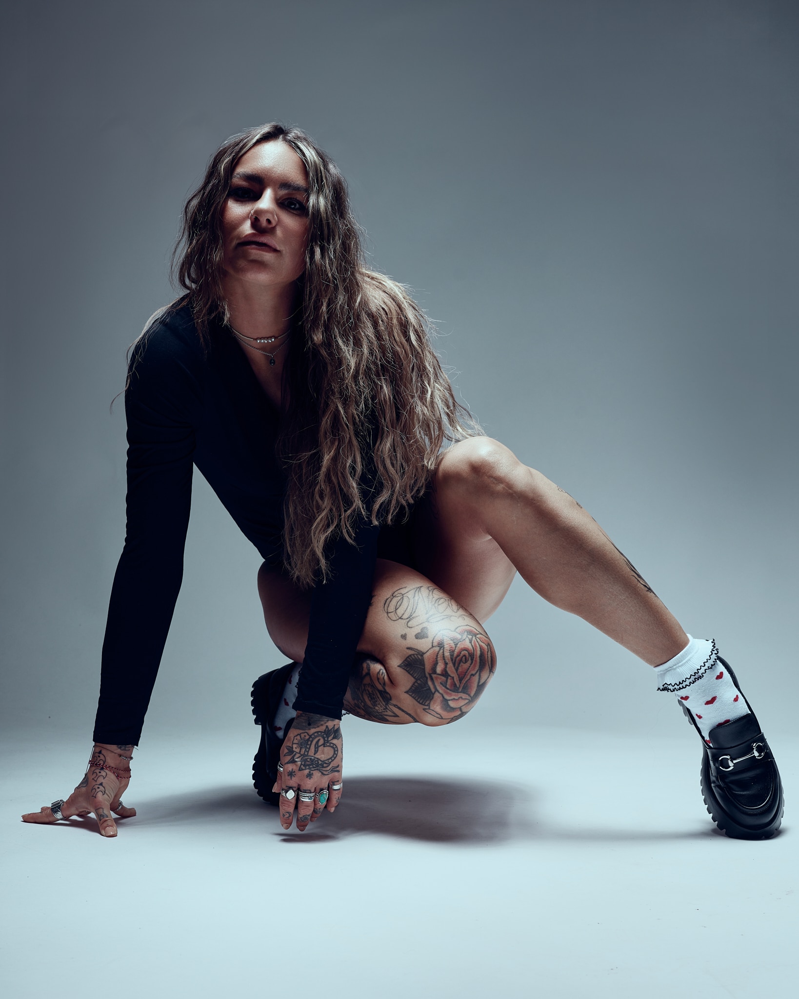 Une femme aux longs cheveux ondulés, tatouée sur les bras et les jambes, vêtue d'un haut noir à manches longues, de chaussures noires et de chaussettes blanches à volants, pose au ras du sol dans un studio à l'arrière-plan neutre.