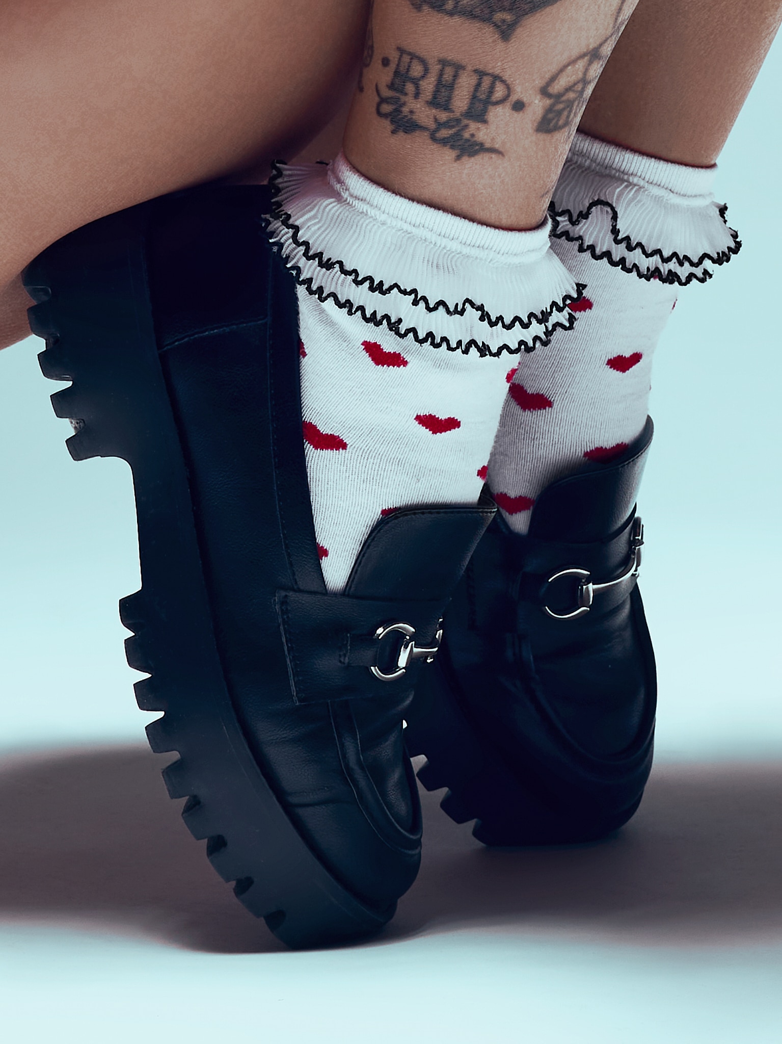 Gros plan sur des jambes portant des chaussettes blanches avec des cœurs rouges et des froufrous noirs, associées à de gros mocassins noirs. Un tatouage indiquant RIP est visible sur une cheville. L'arrière-plan est d'un bleu clair et doux.
