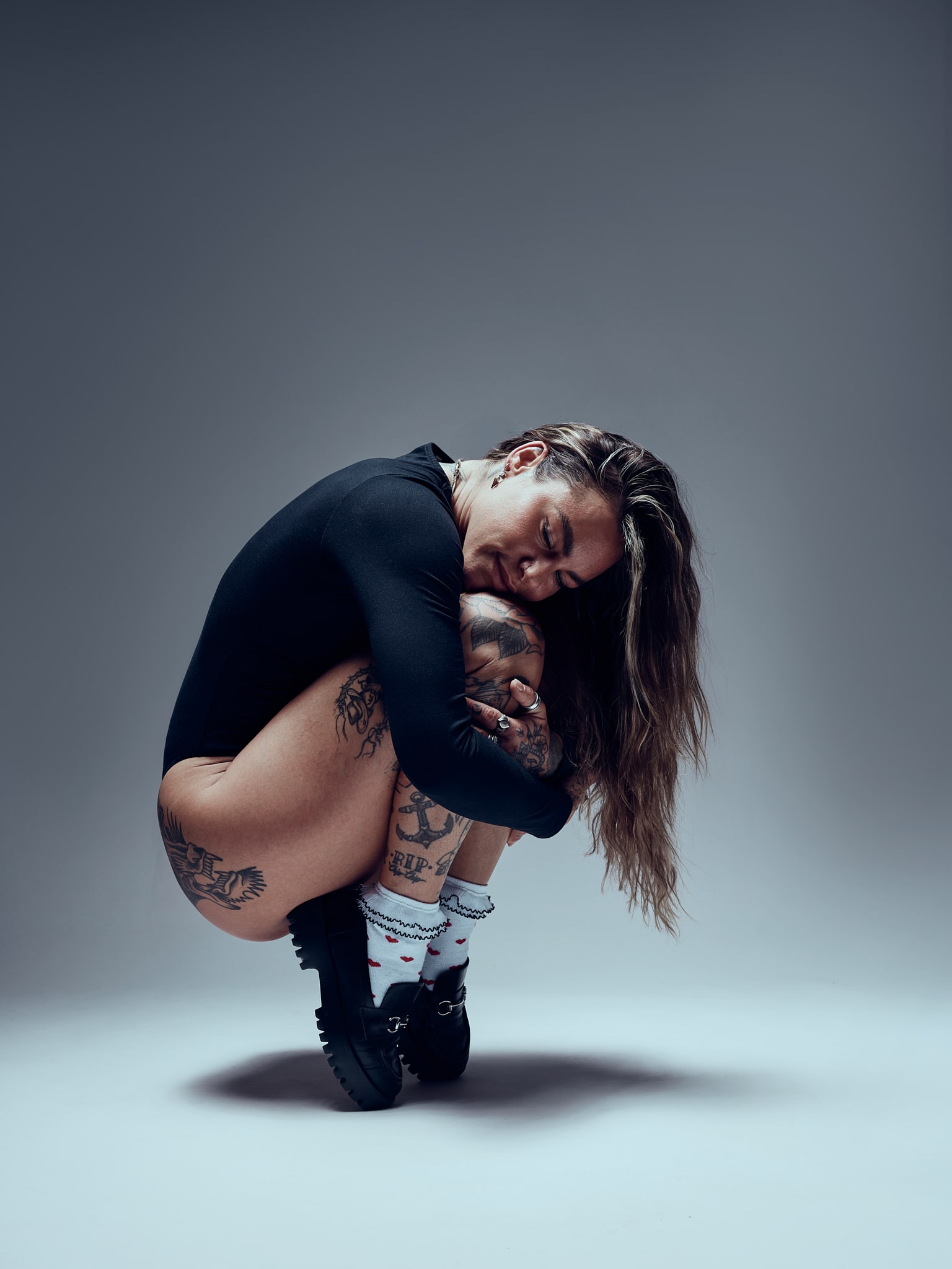 Une personne aux cheveux longs et tatoués est assise en position serrée et recroquevillée sur le sol, serrant ses genoux contre sa poitrine. Elle porte un body noir, des chaussettes à motifs et des chaussures noires, et affiche une expression calme dans un studio.