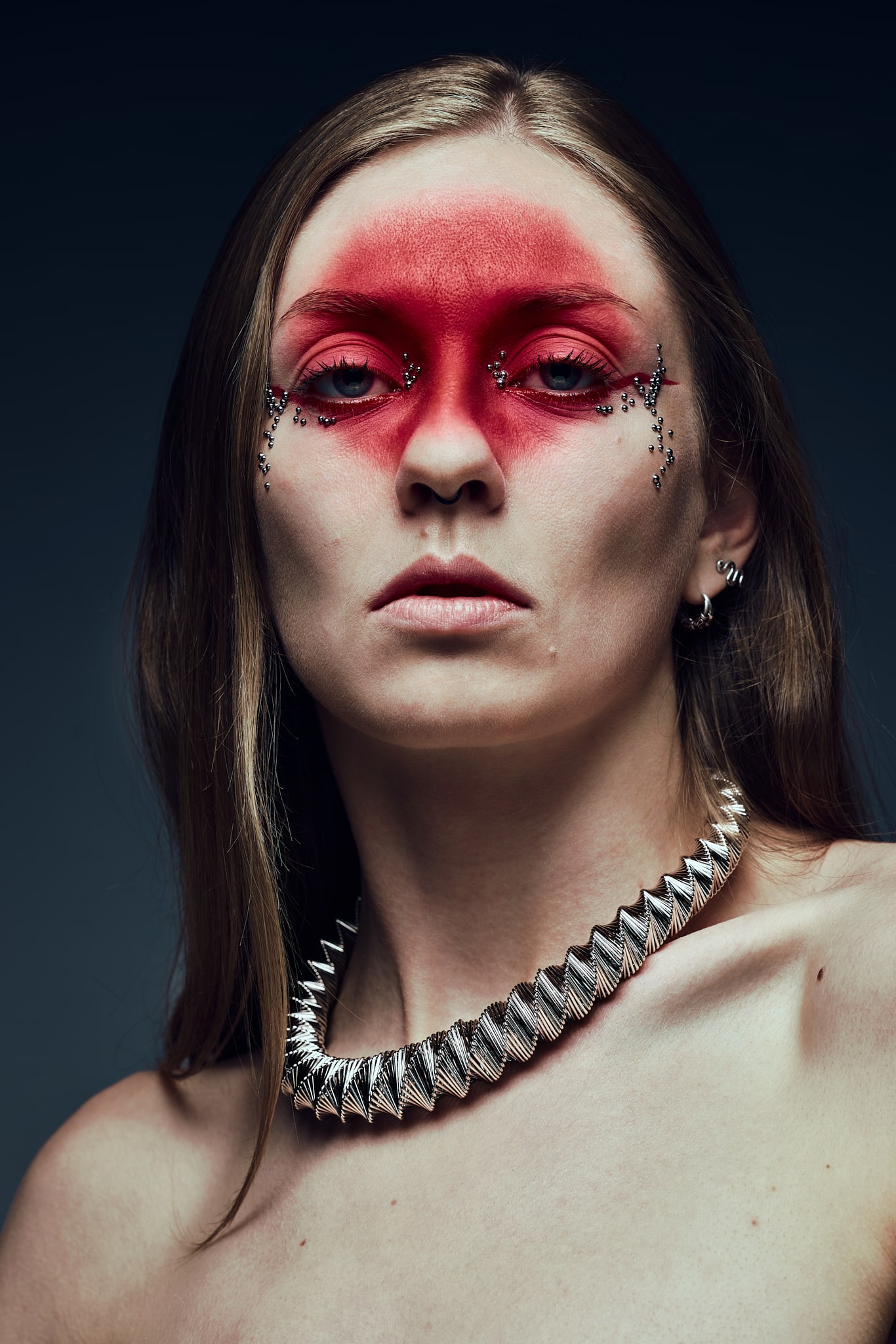 Publicitaire. Une personne aux cheveux longs porte un maquillage rouge spectaculaire autour des yeux, des motifs complexes en strass près des yeux et un collier argenté à pointes. L'arrière-plan est sombre et l'expression est sérieuse.