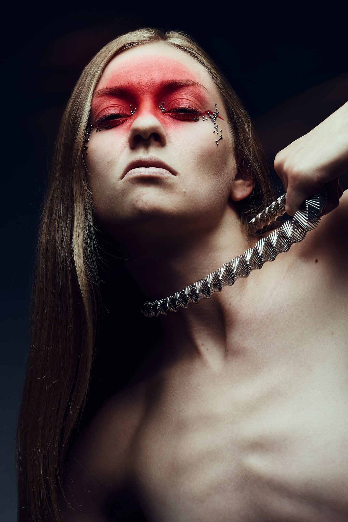 Une personne aux cheveux longs et aux yeux maquillés de rouge fixe intensément l'appareil photo tout en tenant un objet métallique en forme de spirale contre son cou. Son expression est sérieuse et l'arrière-plan est sombre.