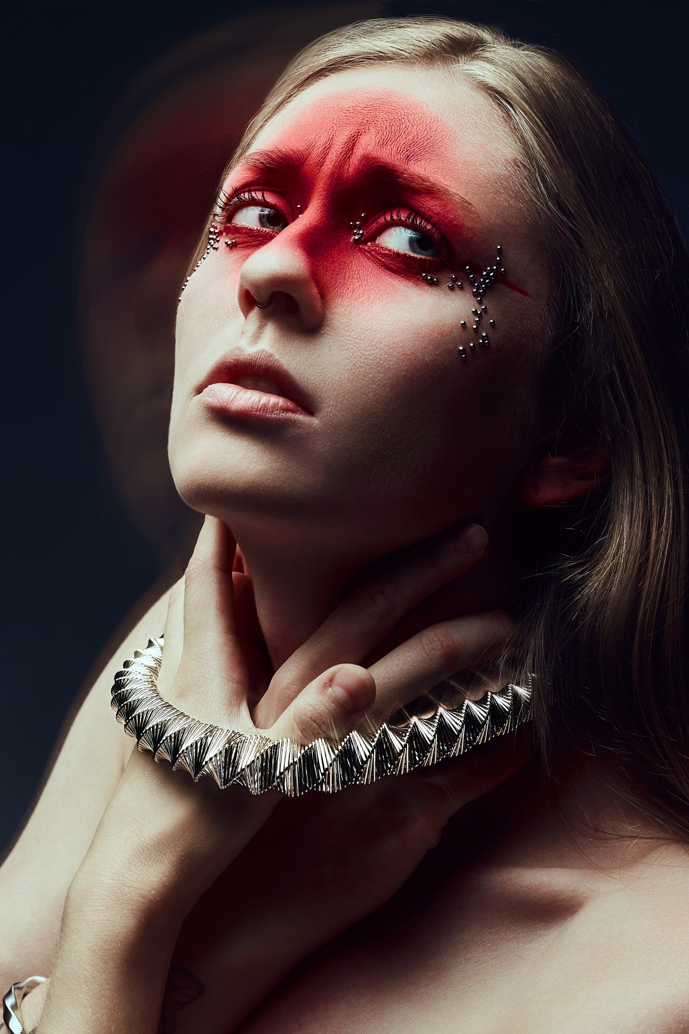 Une personne aux yeux maquillés d'un rouge intense et ornés d'argent regarde fixement vers le haut. Elle porte un collier métallique à pointes et se tient doucement le cou d'une main. L'arrière-plan est sombre, avec un léger effet de double exposition.