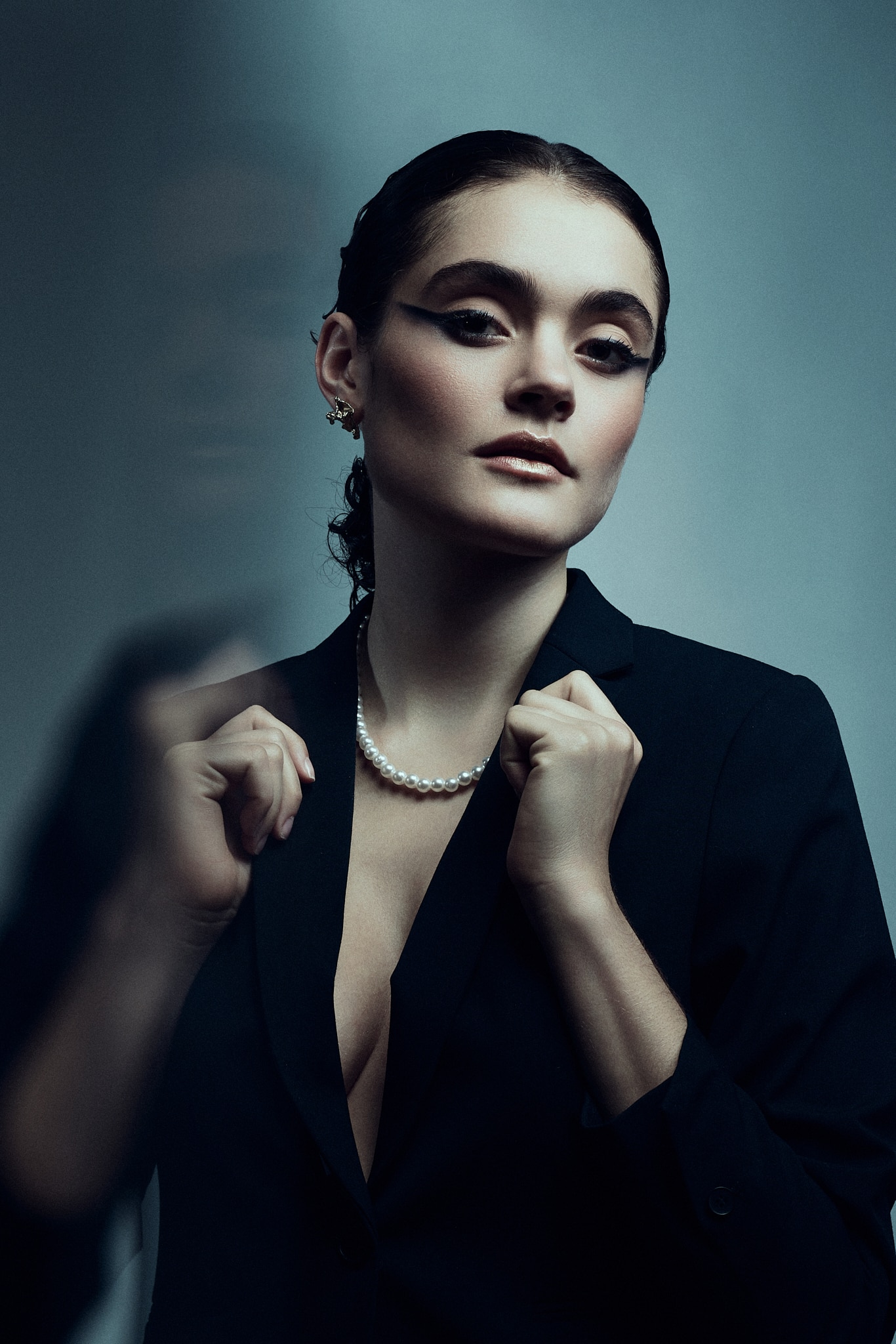 Une femme aux cheveux gominés porte un blazer noir, un collier de perles et un maquillage sombre et audacieux pour les yeux. Elle regarde l'appareil photo avec assurance, tenant les revers de son blazer à deux mains sur un fond bleu-gris.