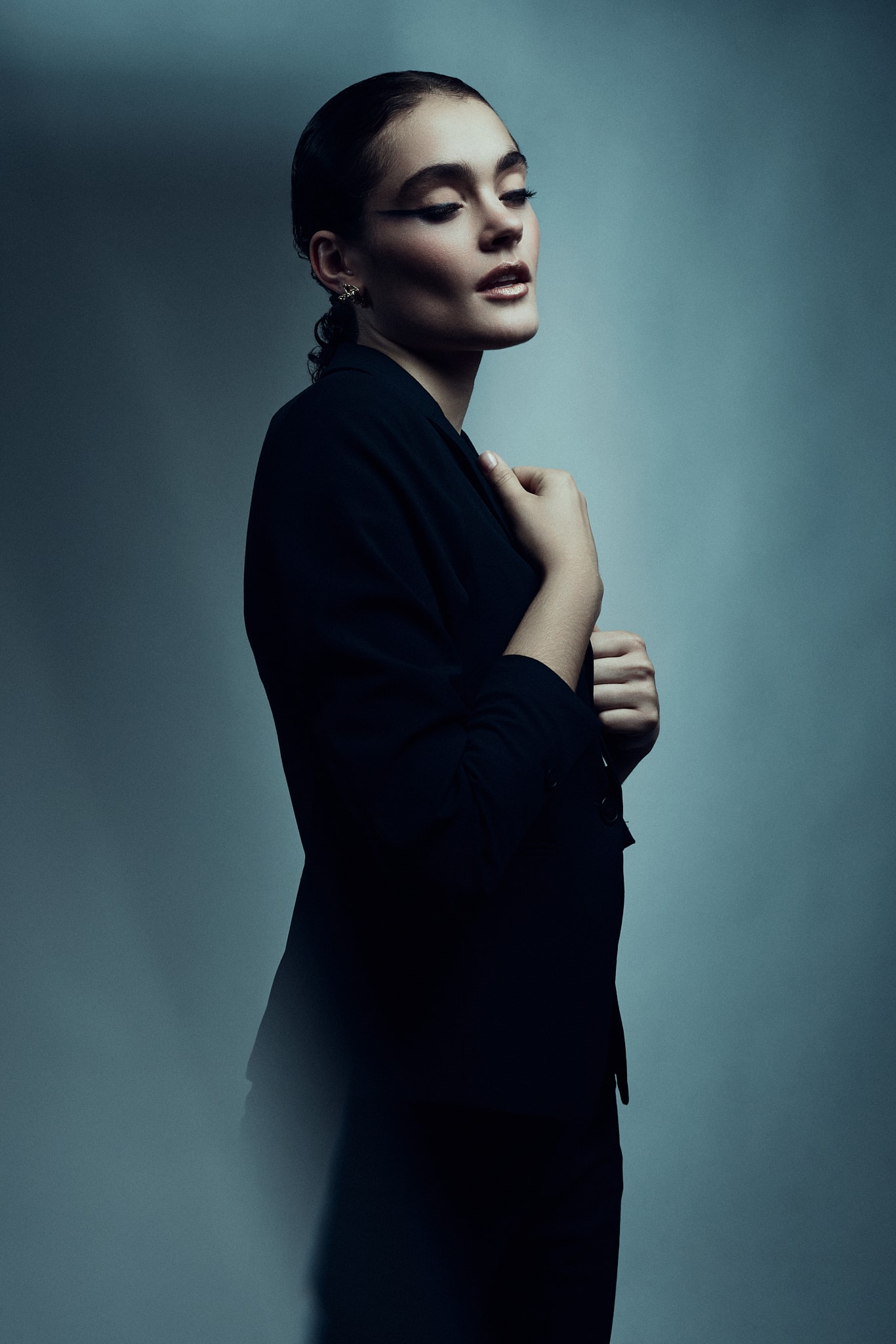 Une personne aux cheveux noirs gominés, vêtue d'un costume noir, se tient debout, les yeux fermés et les mains délicatement serrées contre sa poitrine, sur un fond bleu dégradé, légèrement éclairé.