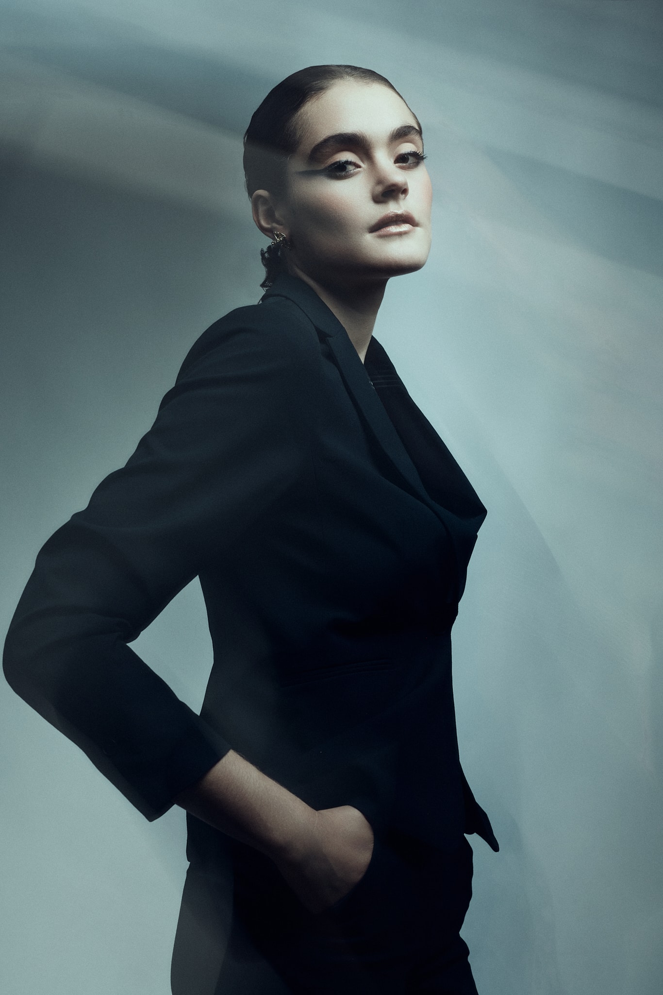 Une personne vêtue d'un costume d'élégance noir se tient debout, les mains dans les poches, et regarde l'appareil photo avec assurance. L'éclairage est sombre et doux, créant une atmosphère dramatique et de haute couture.