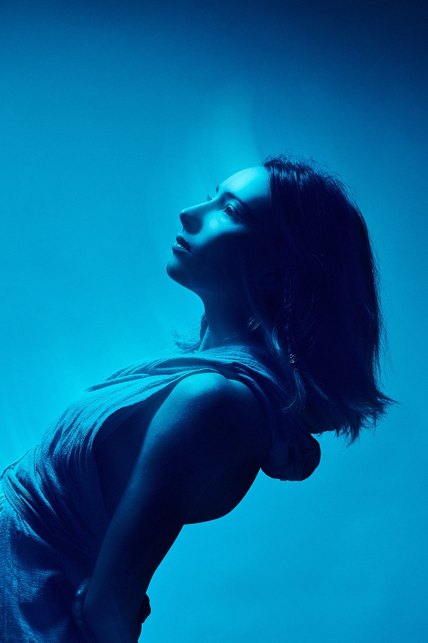 Une femme vêtue d'un haut sans manches pose de profil, penchée en arrière et regardant vers le haut. L'image est éclairée par une lumière bleue dramatique, créant une atmosphère morose et éthérée sur un fond bleu.