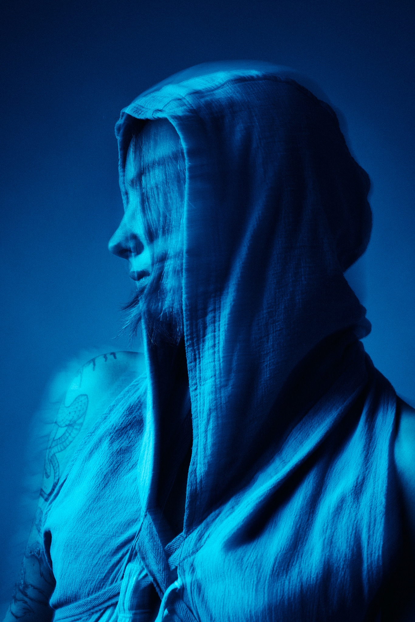 Une personne portant un vêtement à capuche se tient de profil sur un fond bleu, partiellement obscurci par le flou de mouvement et les ombres. L'ensemble de l'image a une teinte bleue, ce qui crée une atmosphère morose et mystérieuse.