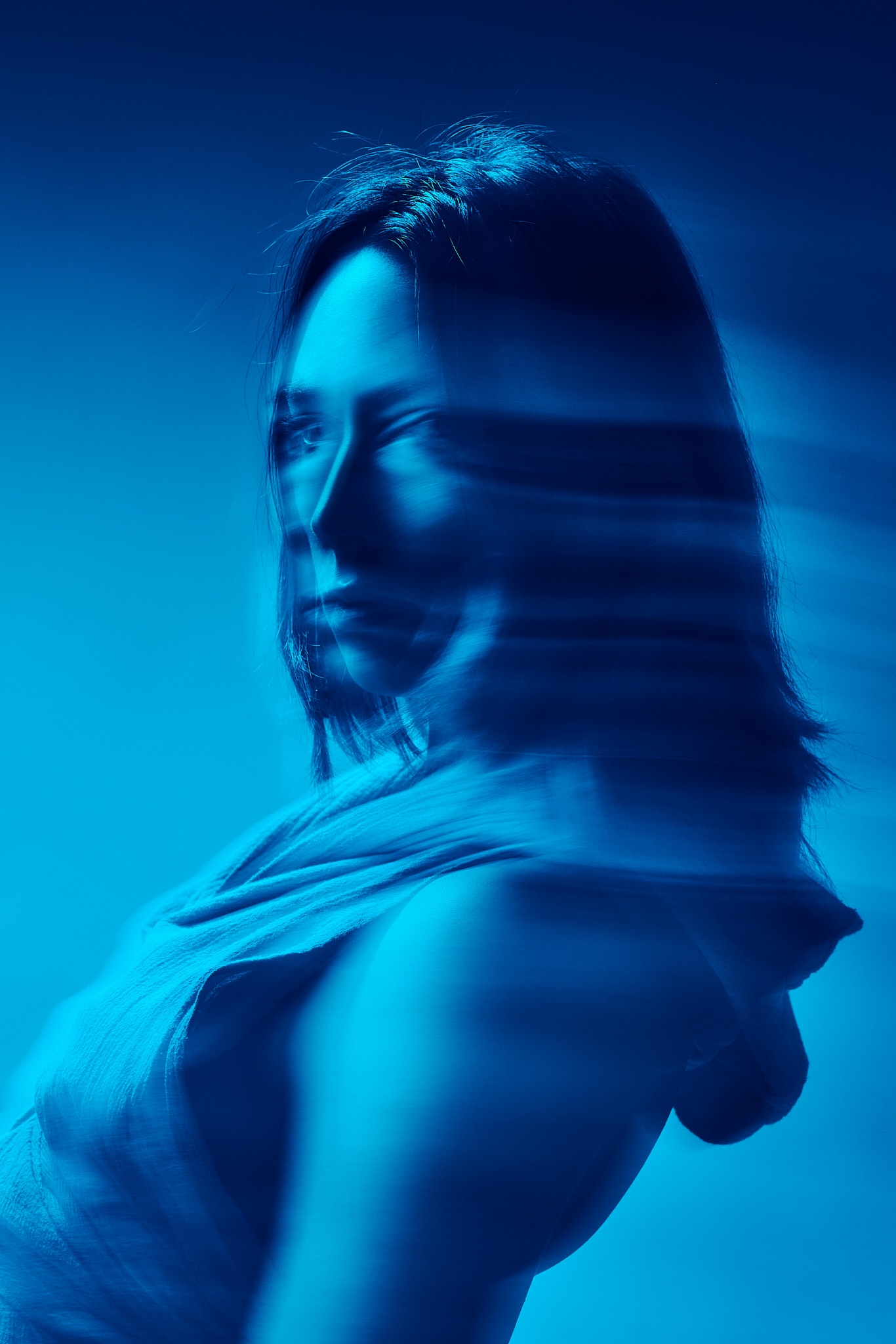 Une personne aux cheveux mi-longs est représentée dans une image teintée de bleu avec un flou de mouvement sur le visage et les épaules, créant un effet onirique et surréaliste. Elle porte un haut sans manches et regarde légèrement par-dessus son épaule.