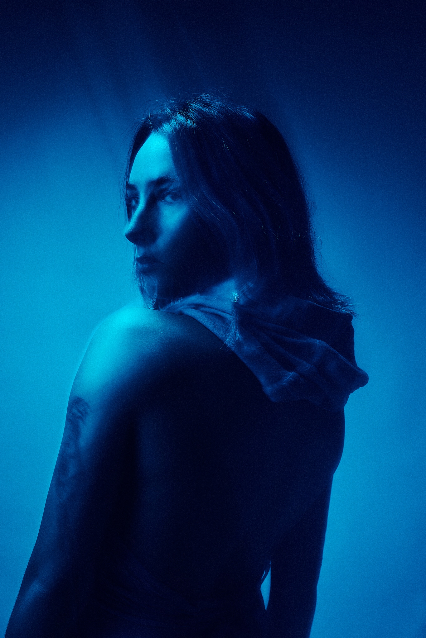 Une personne aux cheveux longs se tient debout, dos à la caméra, regardant par-dessus son épaule dans un éclairage bleu dramatique, créant une atmosphère mystérieuse et éthérée. Un tissu translucide est drapé sur son épaule.