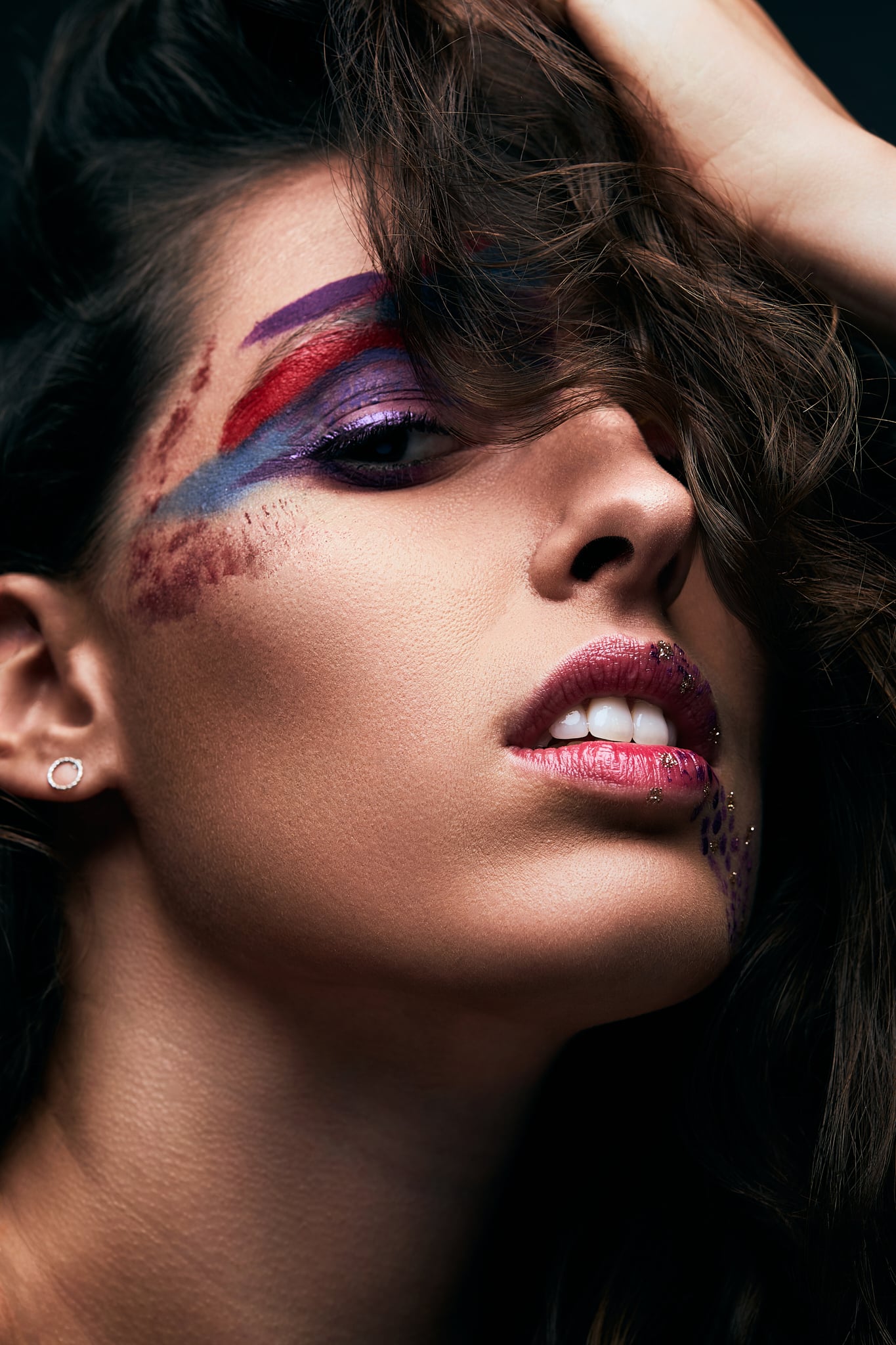 Gros plan sur le visage d'une femme maquillée dans des tons de violet, de bleu et de rouge sur les paupières et les joues, avec un rouge à lèvres texturé. Elle regarde vers le haut, ses cheveux couvrant partiellement son visage.