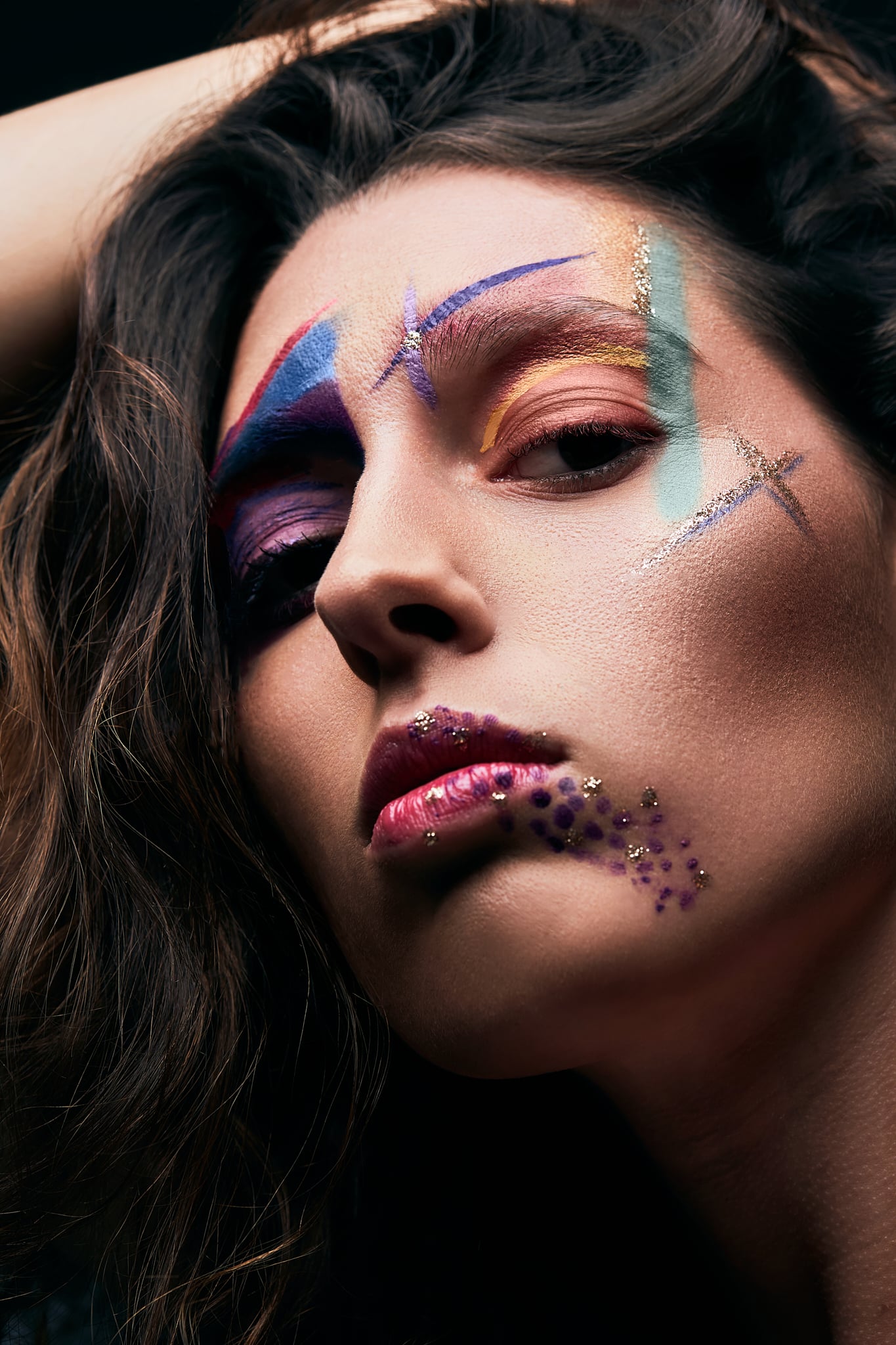 Une femme aux longs cheveux bruns pose, la tête penchée en arrière, avec un maquillage abstrait coloré et audacieux et des paillettes sur le visage et les lèvres, sur un fond sombre.
