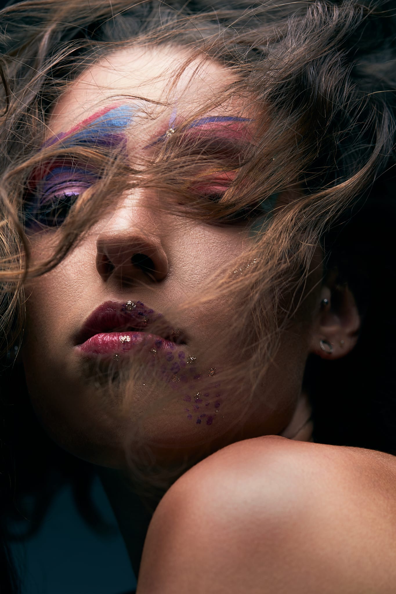 Gros plan graphique sur une personne dont les paupières sont maquillées de couleurs et dont les lèvres et le menton sont pailletés. Des mèches de cheveux cachent partiellement son visage et son épaule est visible au premier plan.