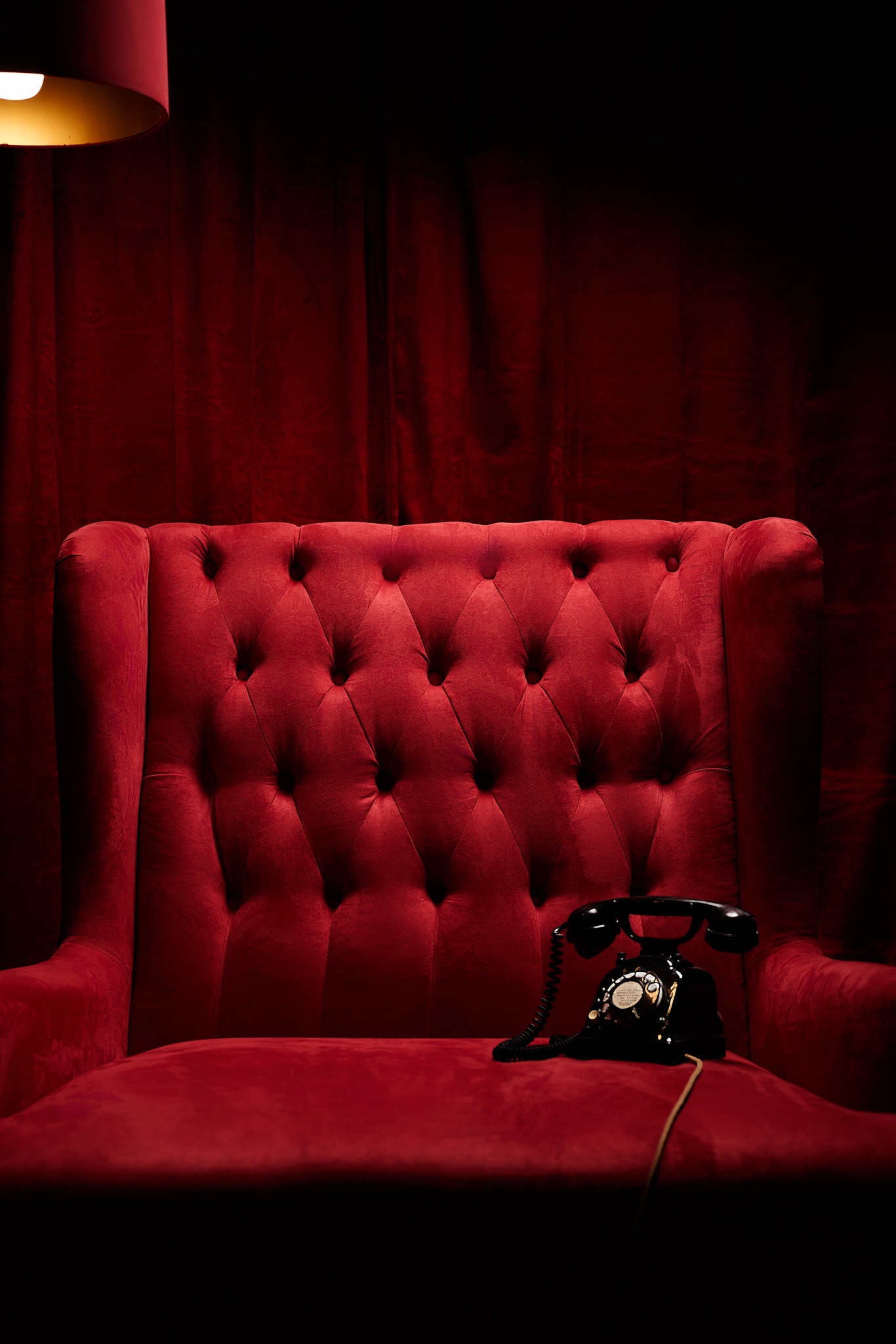 Un fauteuil en velours rouge est assis sous une lampe chaude, avec un téléphone noir à cadran d'époque posé sur son siège. En arrière-plan, de riches rideaux rouges créent une atmosphère dramatique et élégante.