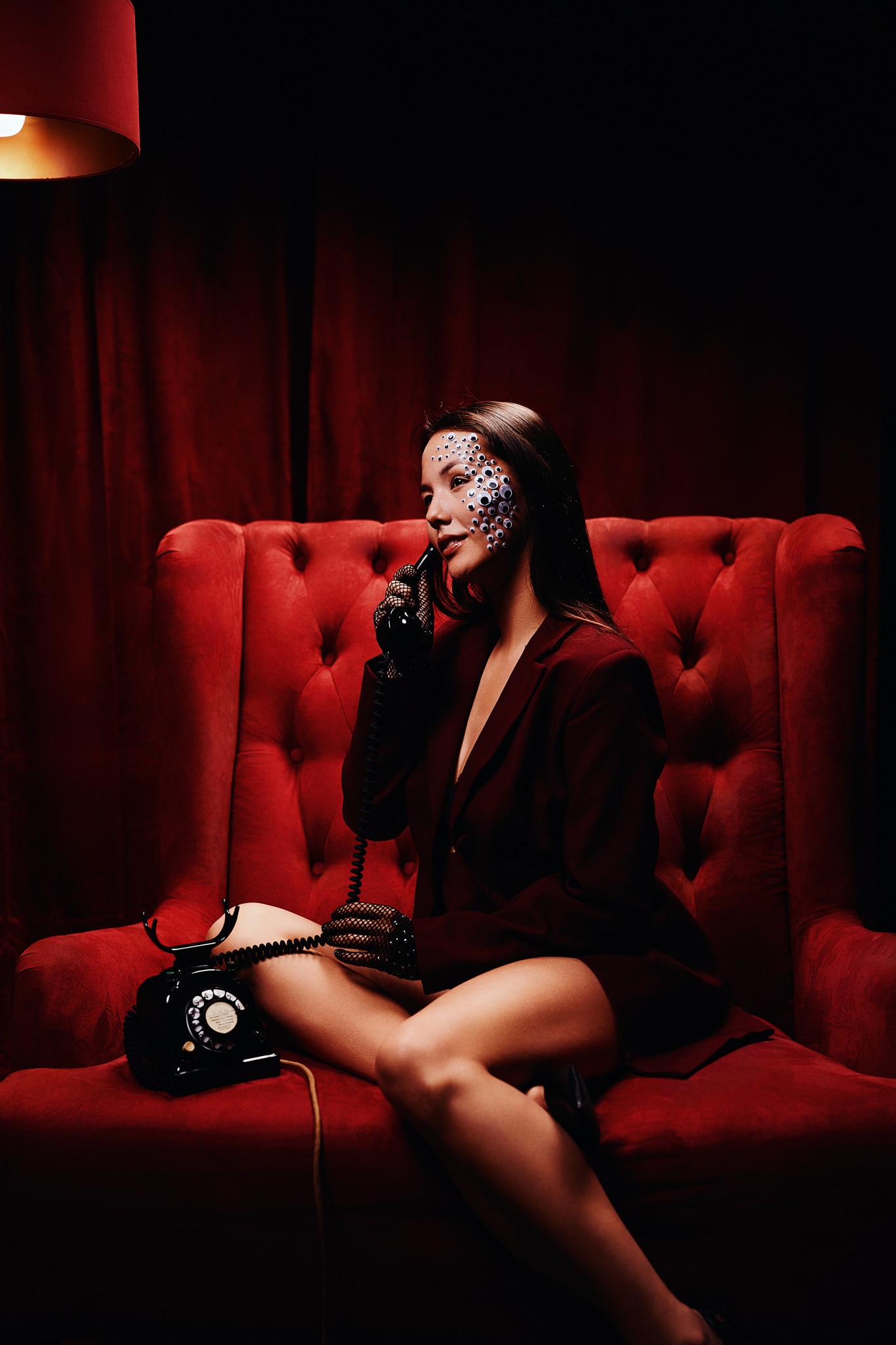 Une femme vêtue d'un blazer sombre est assise sur un fauteuil rouge en peluche et tient un téléphone à cadran à l'oreille. Elle porte des paillettes décoratives sur un côté de son visage et des gants noirs. L'arrière-plan et l'éclairage sont d'un rouge profond.