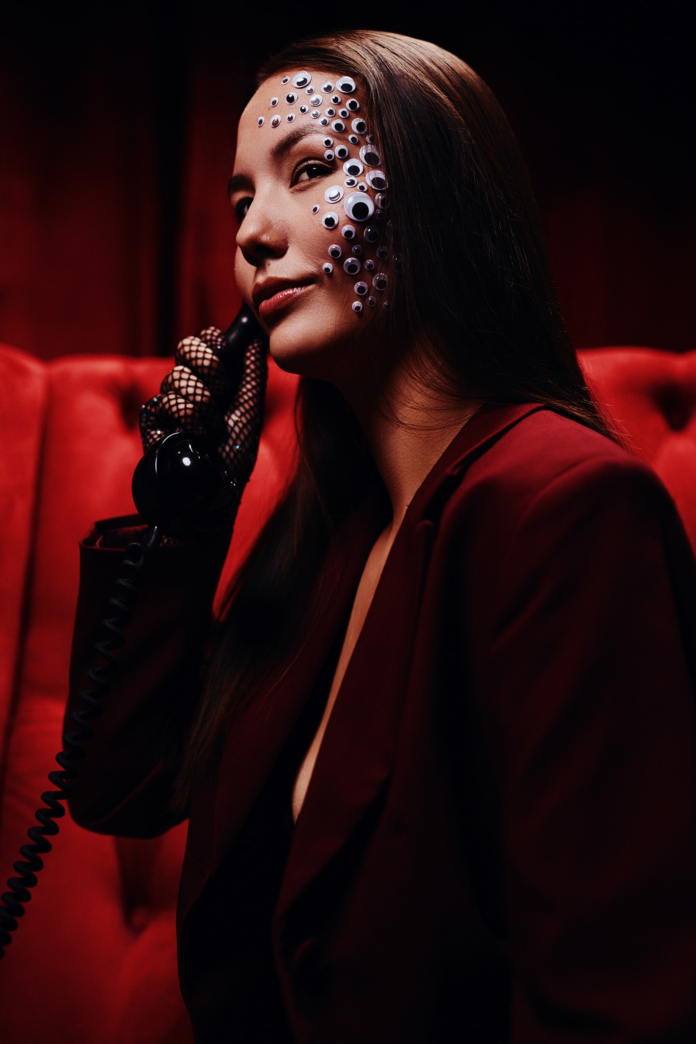 Une femme vêtue d'un blazer rouge foncé est assise sur un canapé touffu rouge, tenant un téléphone noir d'époque. Elle a de longs cheveux bruns et porte des gants en résille noirs. Son visage est orné de multiples petits yeux de poule.