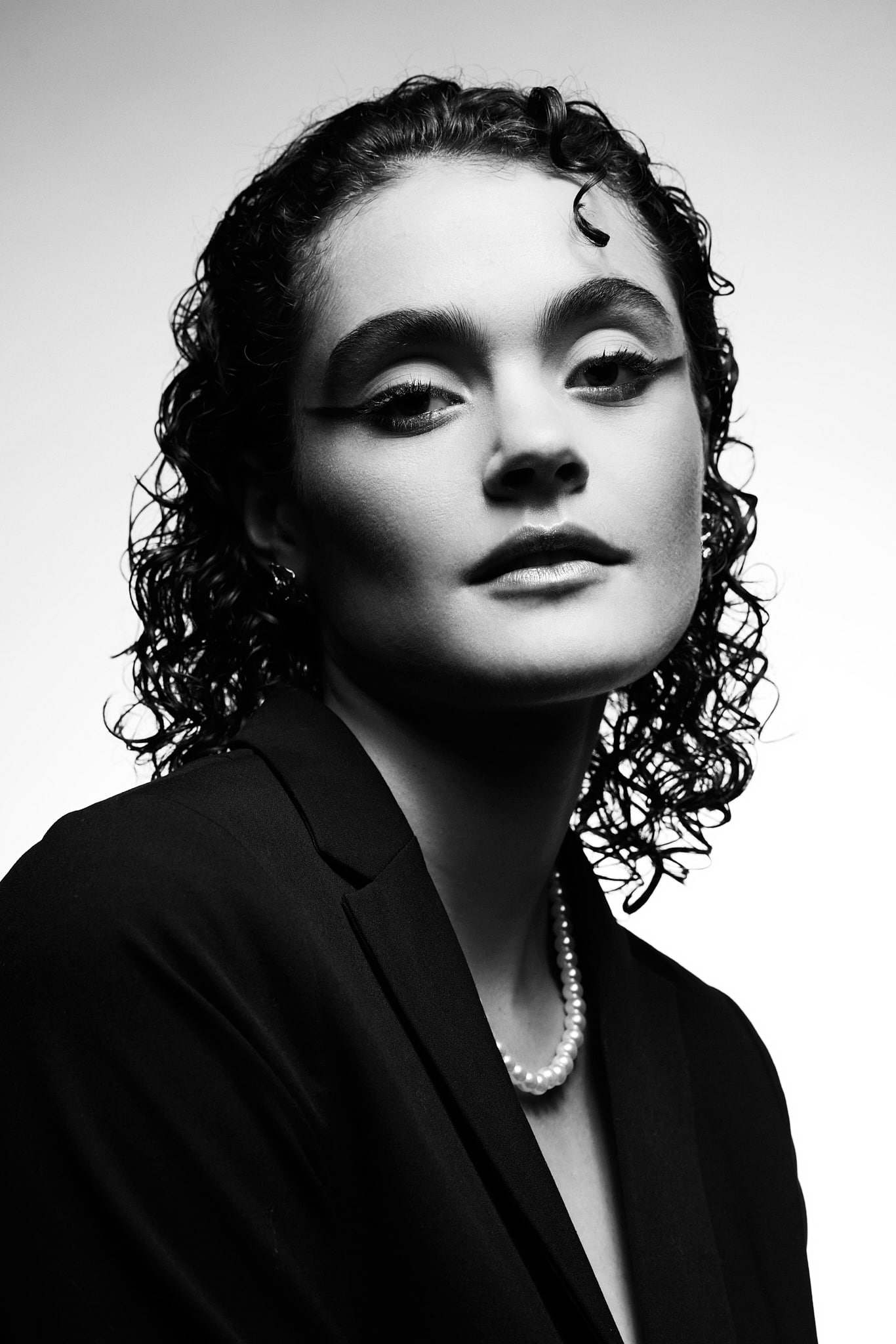 Portrait en noir et blanc d'une personne aux cheveux courts et bouclés, portant un blazer sombre, un collier de perles et un eye-liner audacieux. L'éclairage met en valeur les traits du visage sur un fond uni.