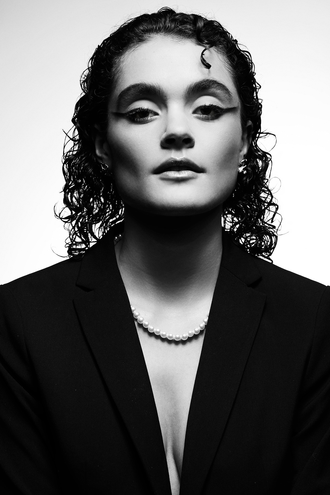 Une femme aux cheveux mouillés et bouclés porte un blazer noir, un collier de perles et un maquillage des yeux audacieux. Elle regarde l'appareil photo avec confiance et une expression neutre. La photo est en noir et blanc avec un éclairage dramatique.