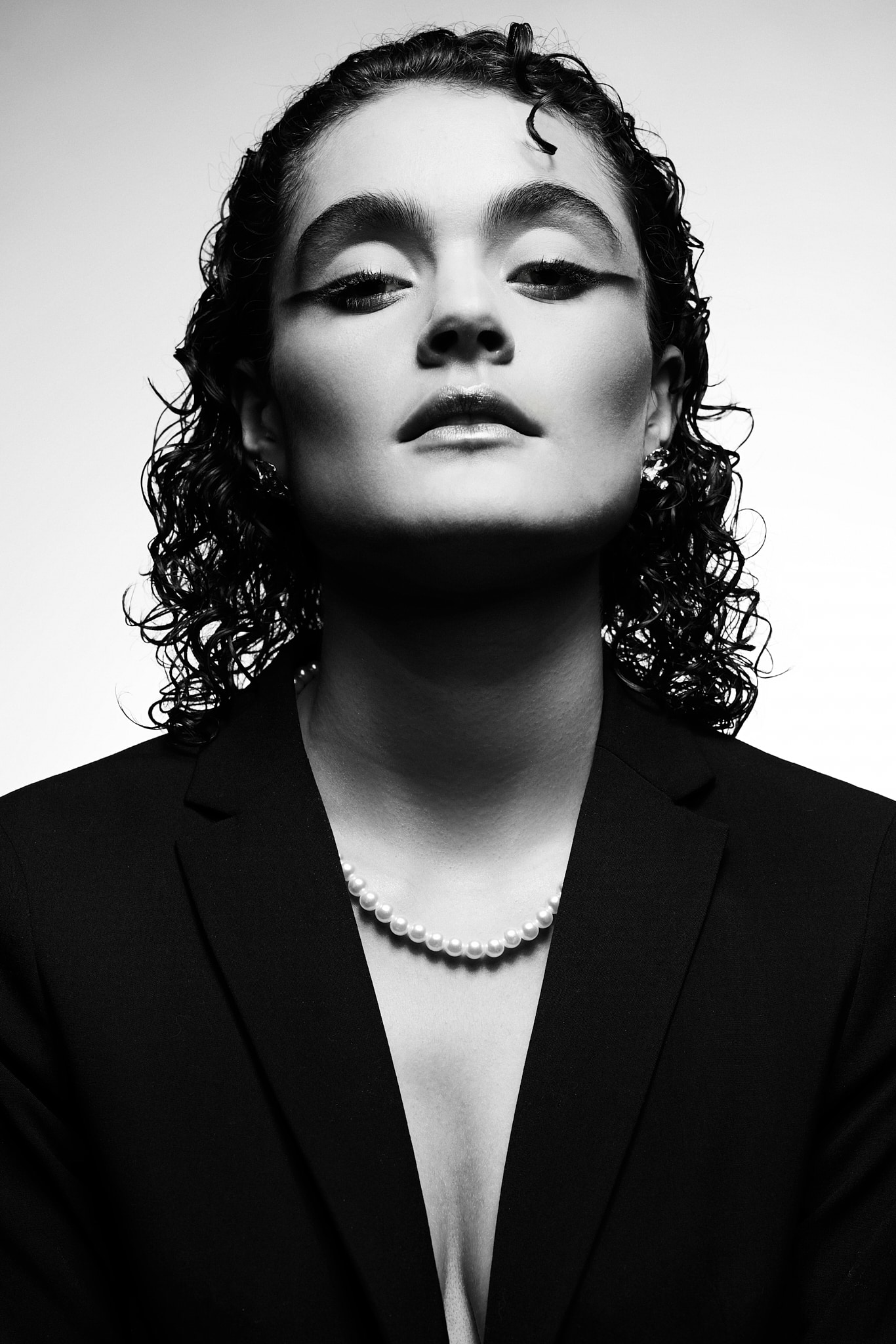 Book agence. Portrait en noir et blanc d'une personne aux cheveux bouclés, aux yeux maquillés et à l'expression confiante, portant un blazer sombre et un collier de perles, sur un fond clair uni.