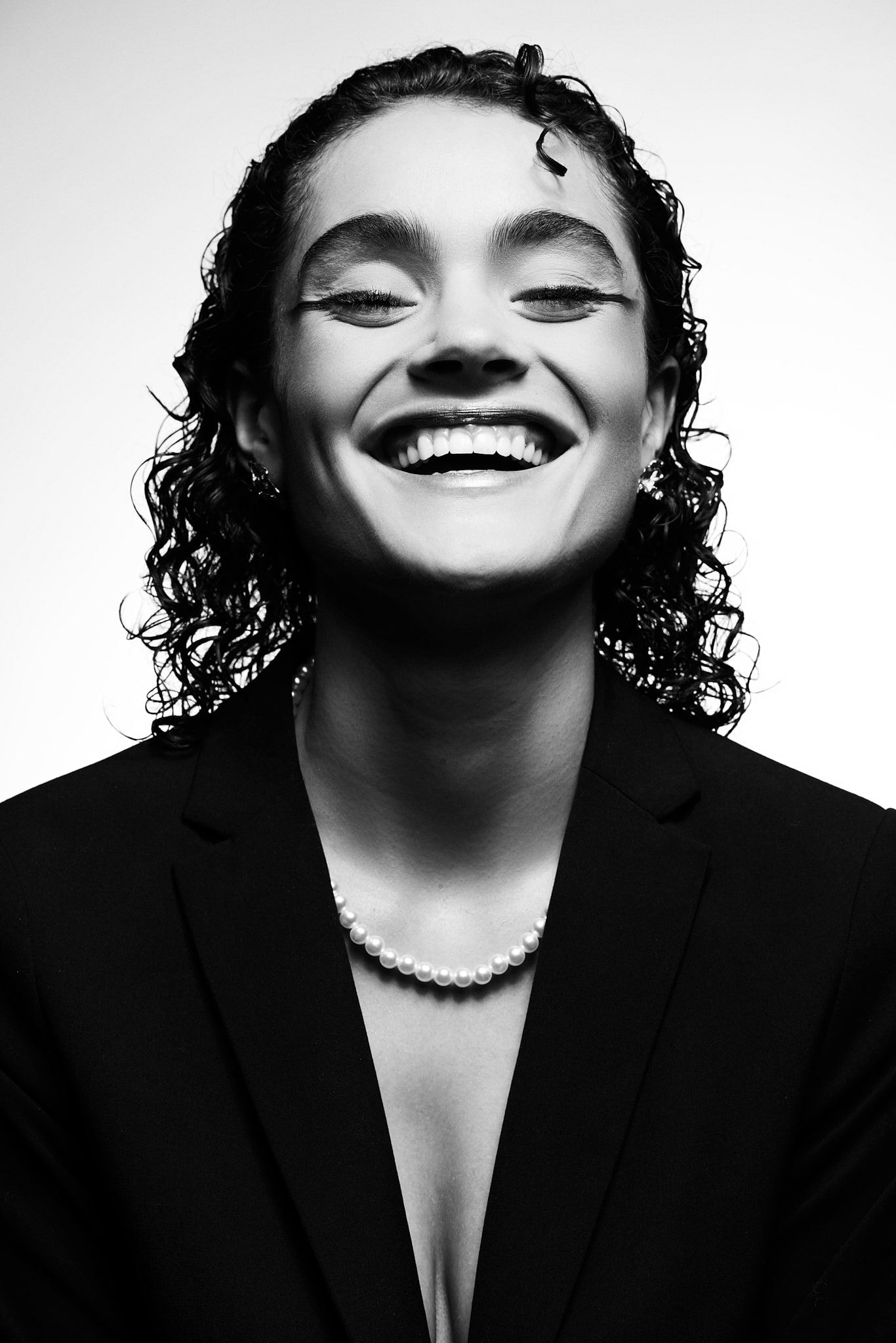 Portrait en noir et blanc d'une personne souriante aux cheveux bouclés, portant un blazer sombre, un collier de perles et un maquillage des yeux audacieux, regardant l'appareil photo avec confiance sur un fond uni.