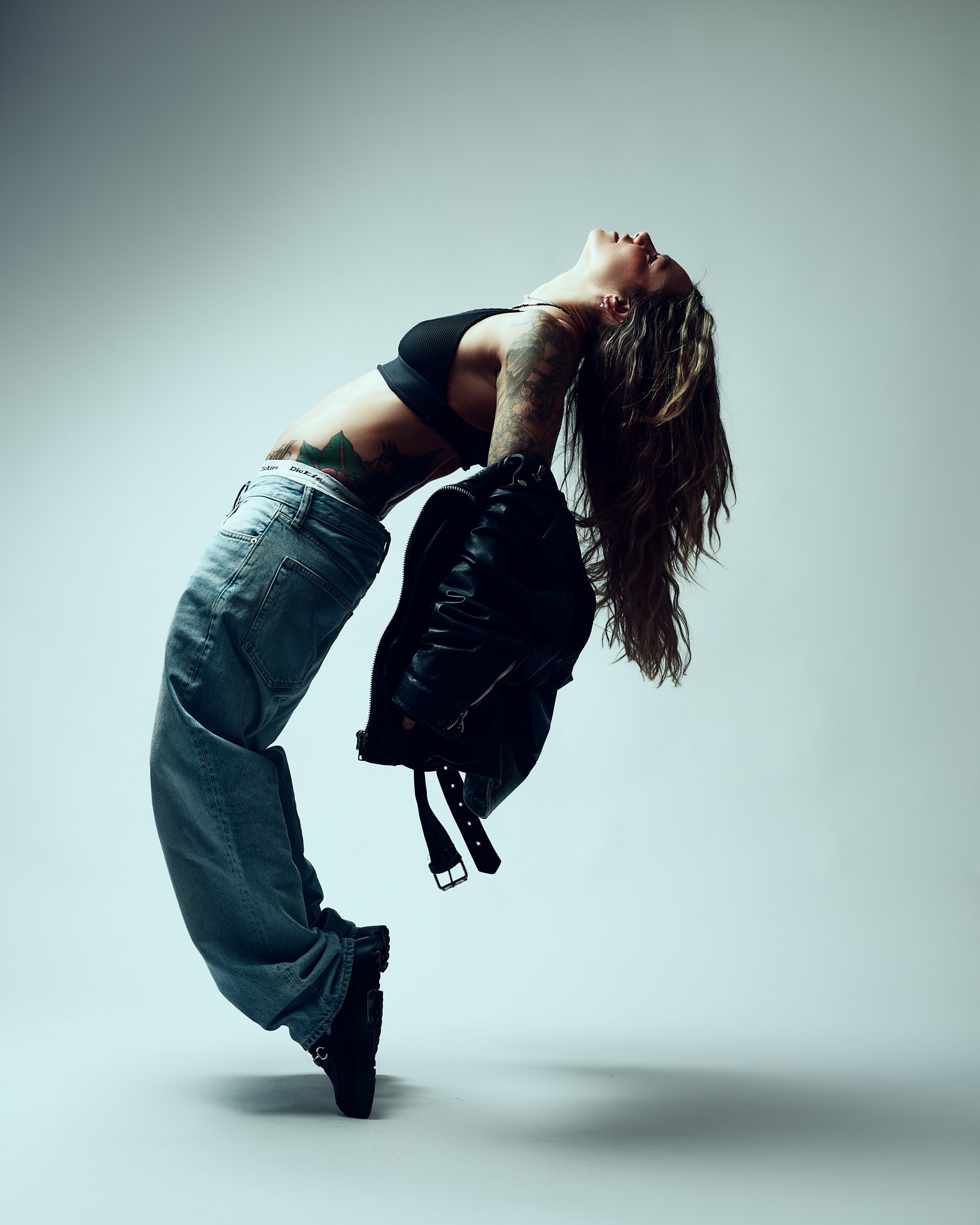 Une personne aux cheveux longs et tatoués, vêtue d'un crop top noir et d'un jean ample, se penche en arrière dans une pose dramatique tout en tenant une veste noire, sur un fond clair uni.