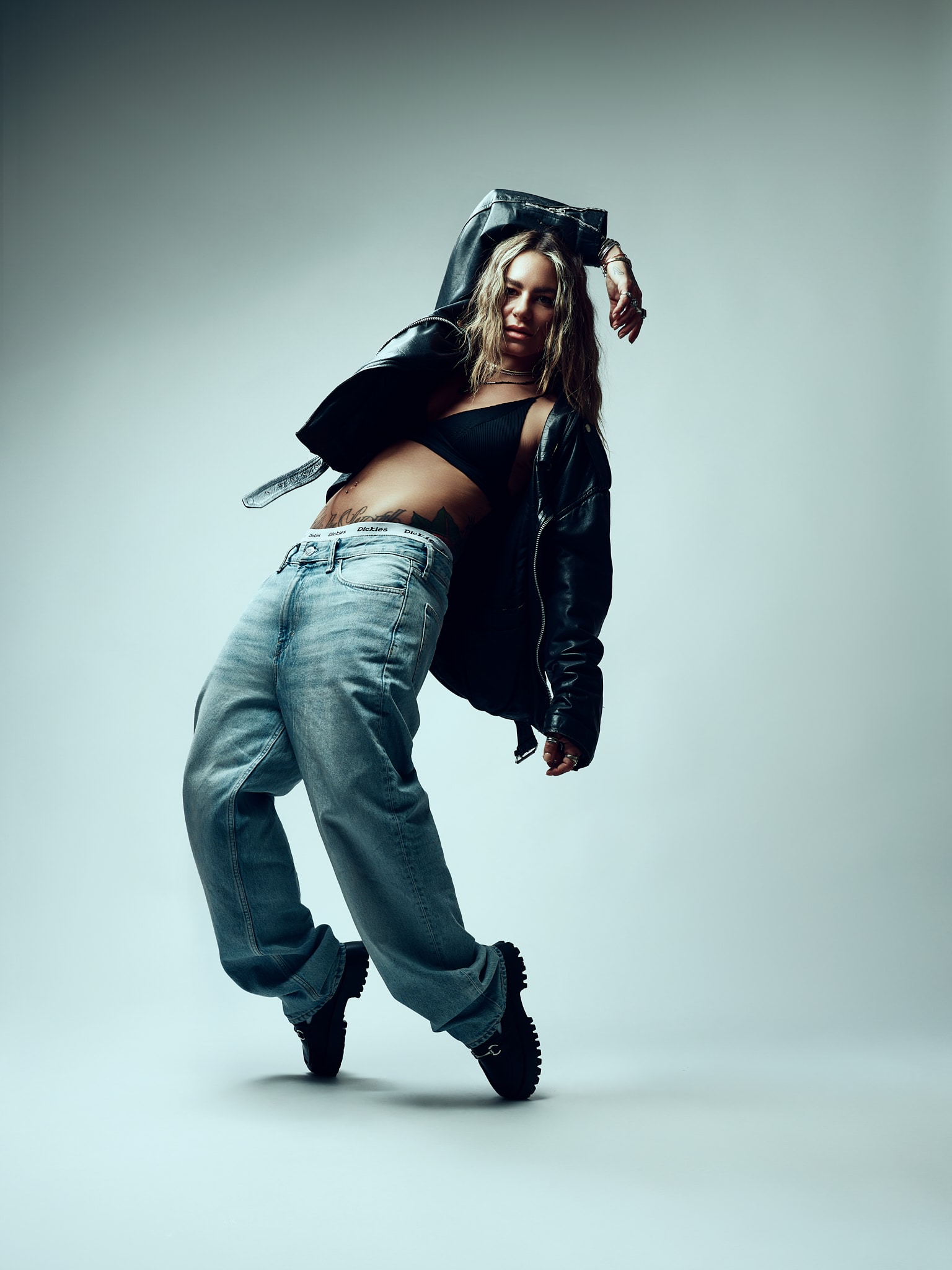 Chorégraphe. Une femme pose de manière dynamique sur un fond uni, vêtue d'un haut noir, d'une veste en cuir noir ample, d'un jean bleu clair ample et de chaussures noires. Son corps est penché sur le côté, un bras levé au-dessus de sa tête.