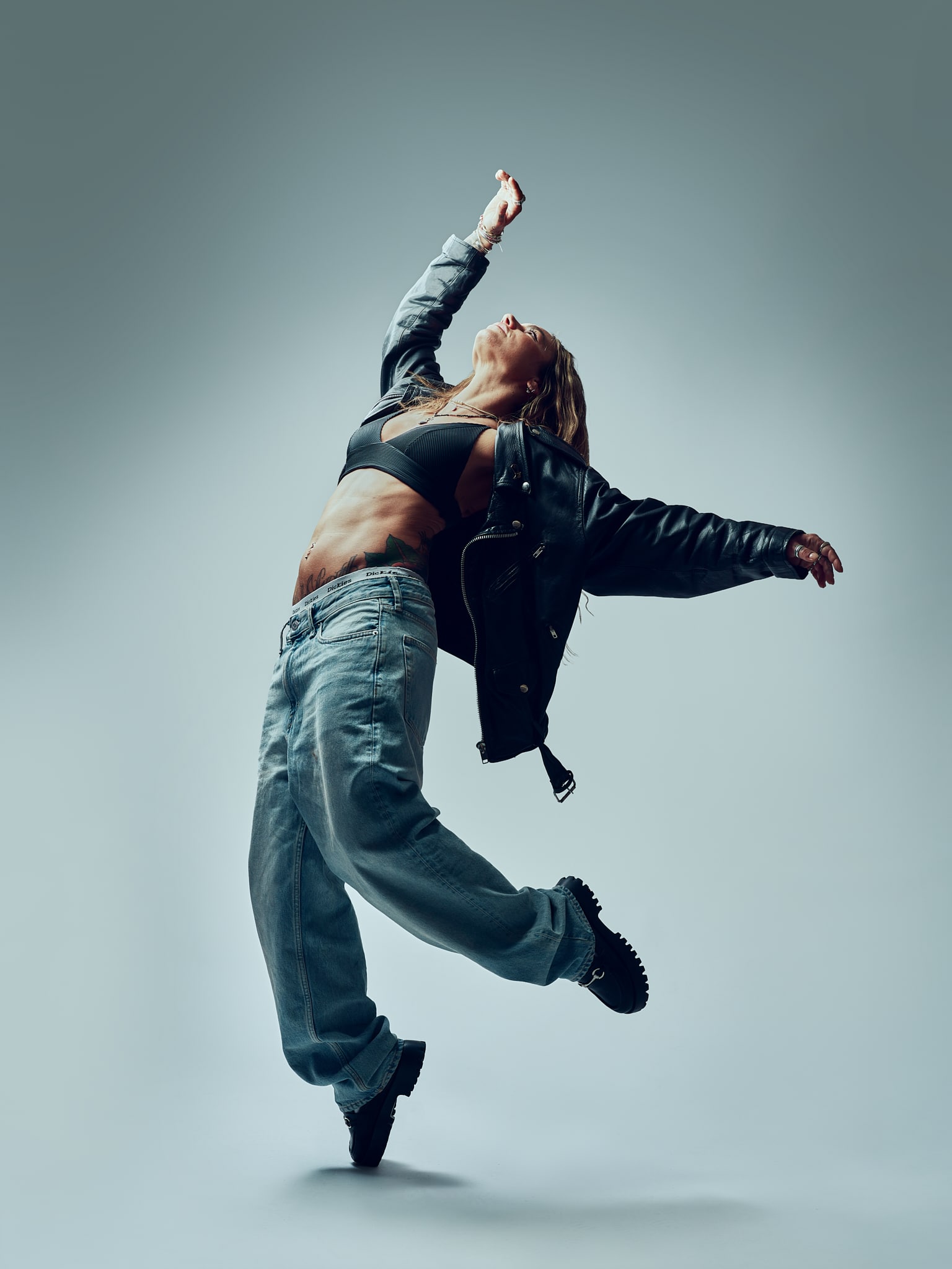 Une femme vêtue d'un jean ample, d'un soutien-gorge noir et d'une veste noire prend une pose de danse dynamique, en équilibre sur une jambe, les bras écartés et la tête inclinée vers l'arrière, sur un fond uni.