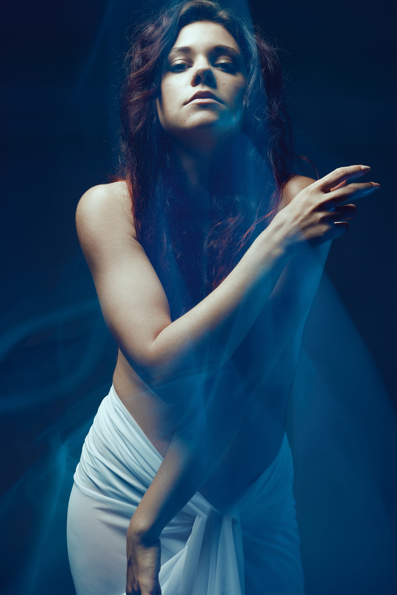 Une femme aux longs cheveux noirs pose avec assurance, drapée dans un tissu blanc sur un fond sombre, avec de la fumée bleue tourbillonnant autour d'elle.