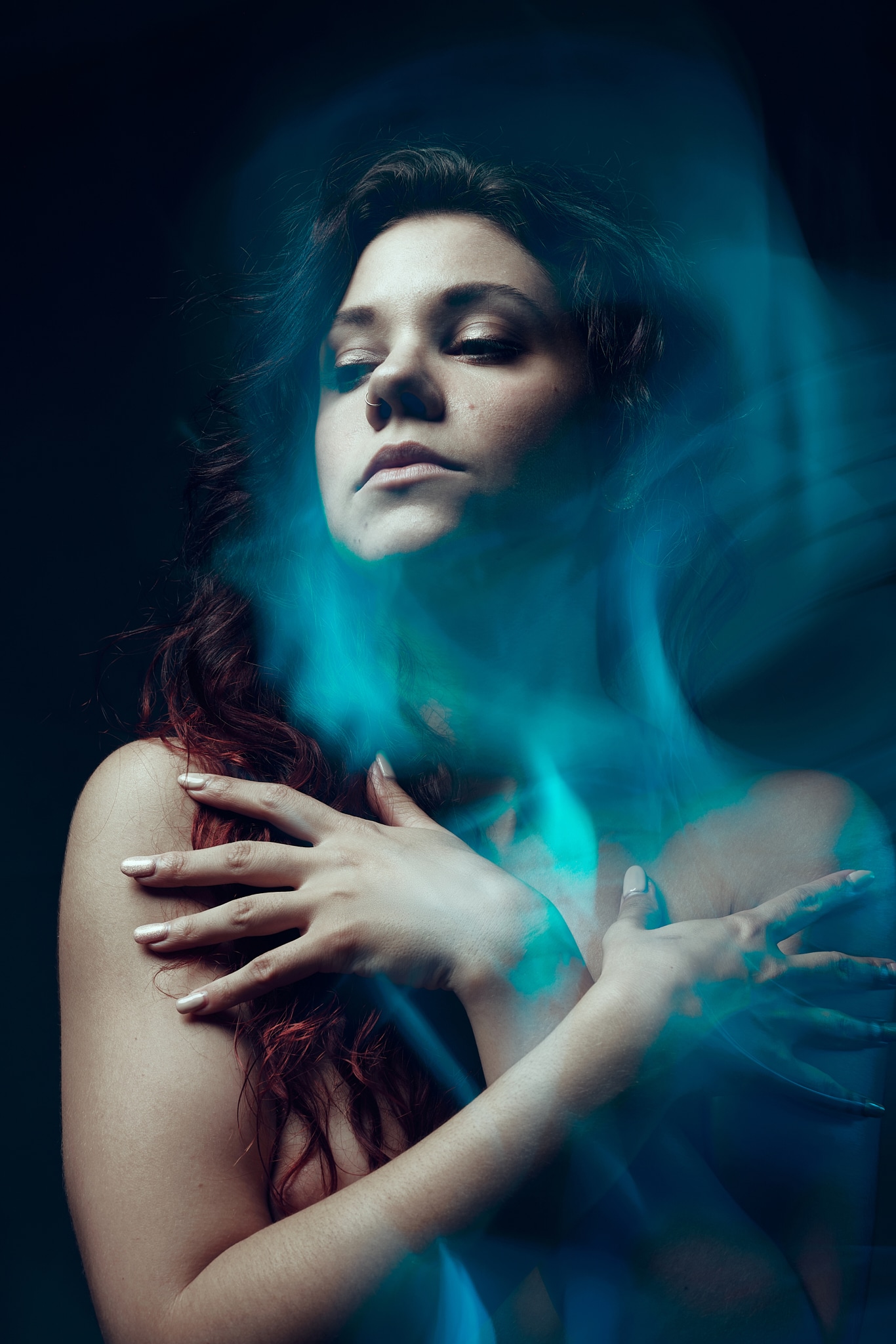 Une femme aux longs cheveux noirs croise les bras sur sa poitrine, les yeux fermés, entourée de traînées lumineuses bleues éthérées dans un décor sombre et morose.