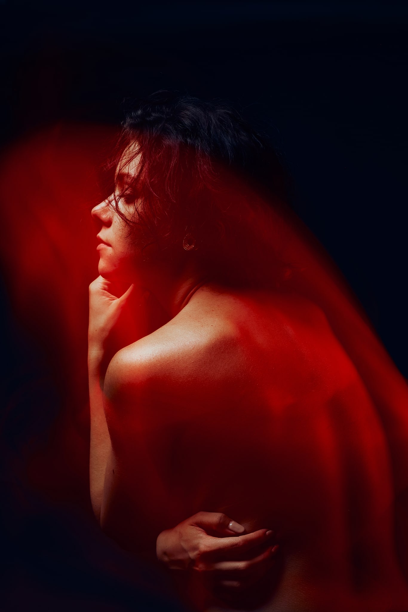 Une personne aux épaules dénudées pose de profil sur un fond sombre, éclairé par des effets de lumière dramatique rouge et douce en mouvement autour d'elle, créant une atmosphère sombre et artistique.