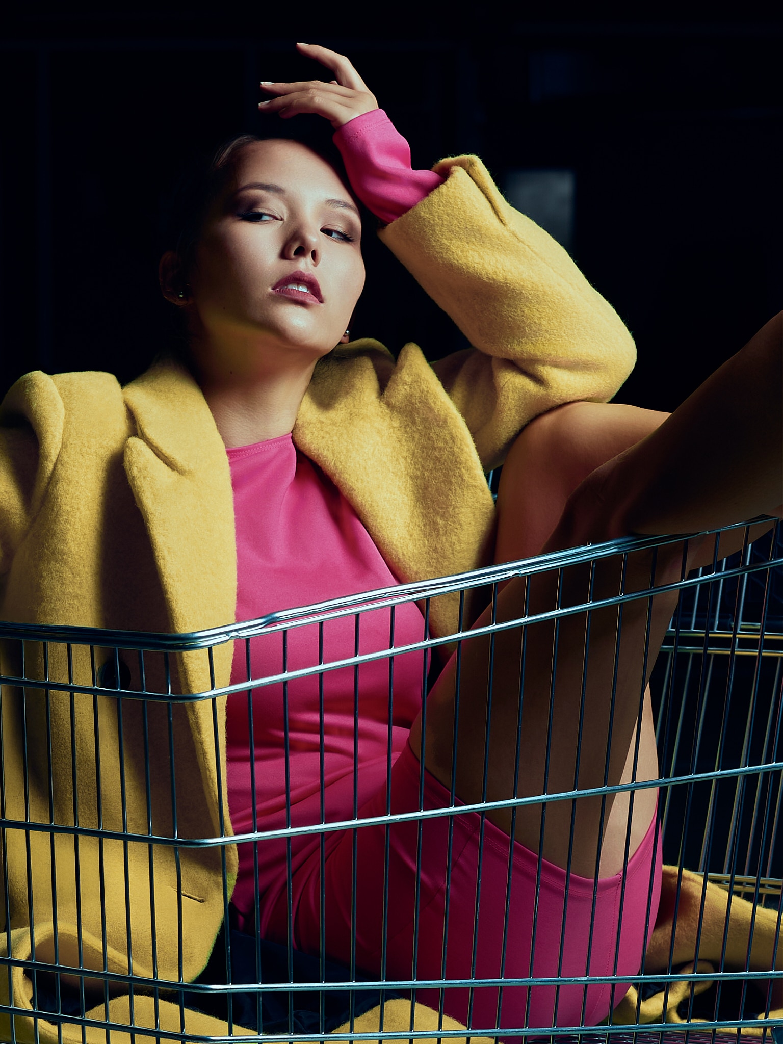 Une femme vêtue d'un manteau jaune et d'une tenue rose pose allongée dans un chariot de supermarché en métal, une main sur la tête et une jambe levée, sur un fond sombre.